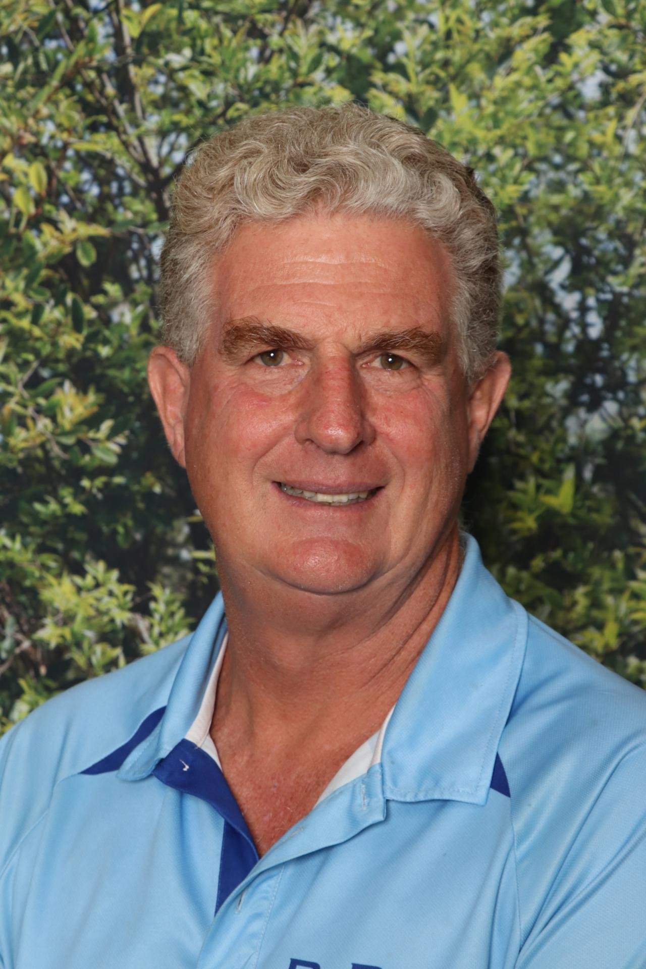 A middle-aged man with gray curly hair and light skin smiling, wearing a light blue collared shirt against a background of green leafy trees.