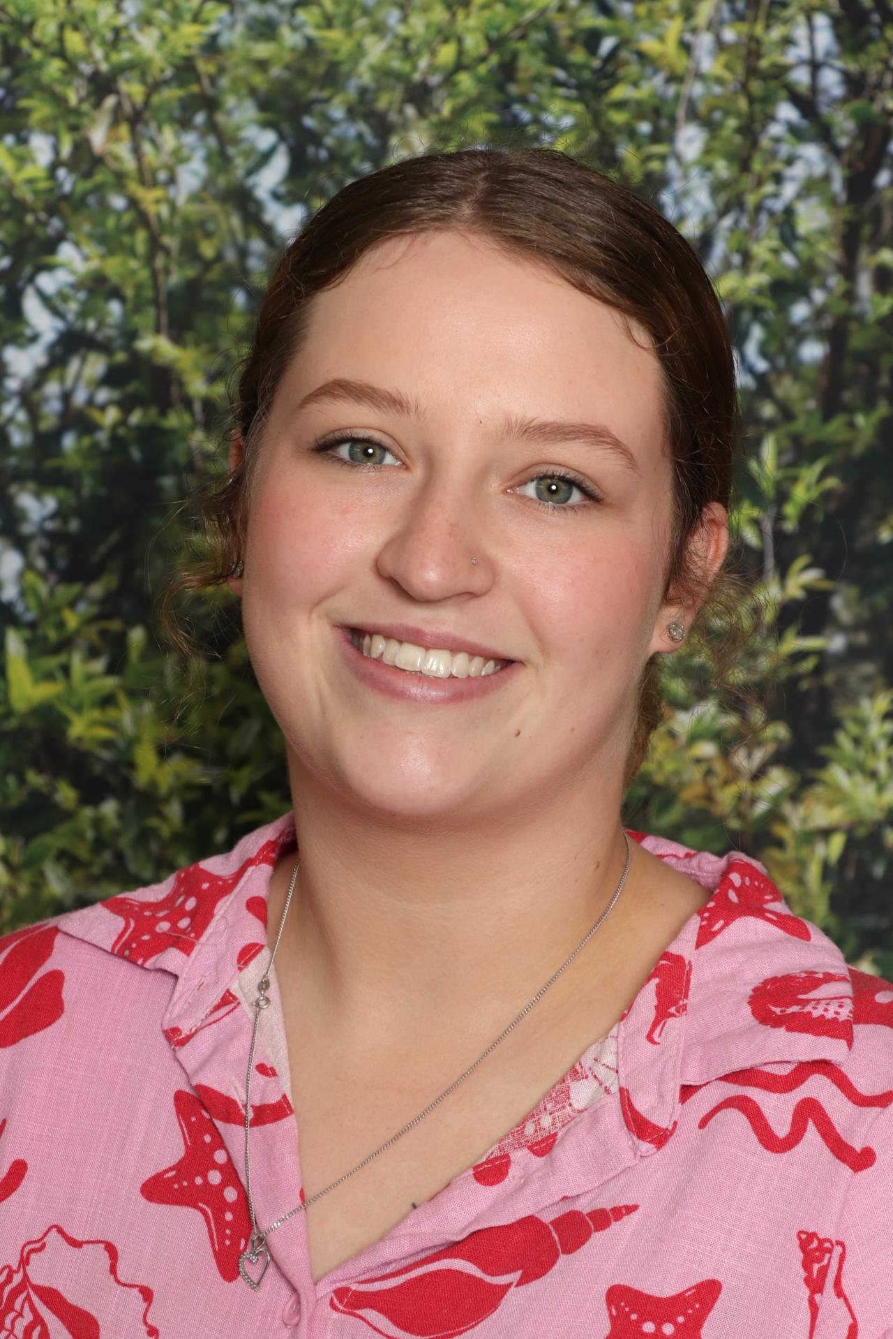 Young woman smiling with blue eyes and brown hair tied back, wearing a pink shirt with red lobster and starfish print, standing outdoors with blurred green foliage background.