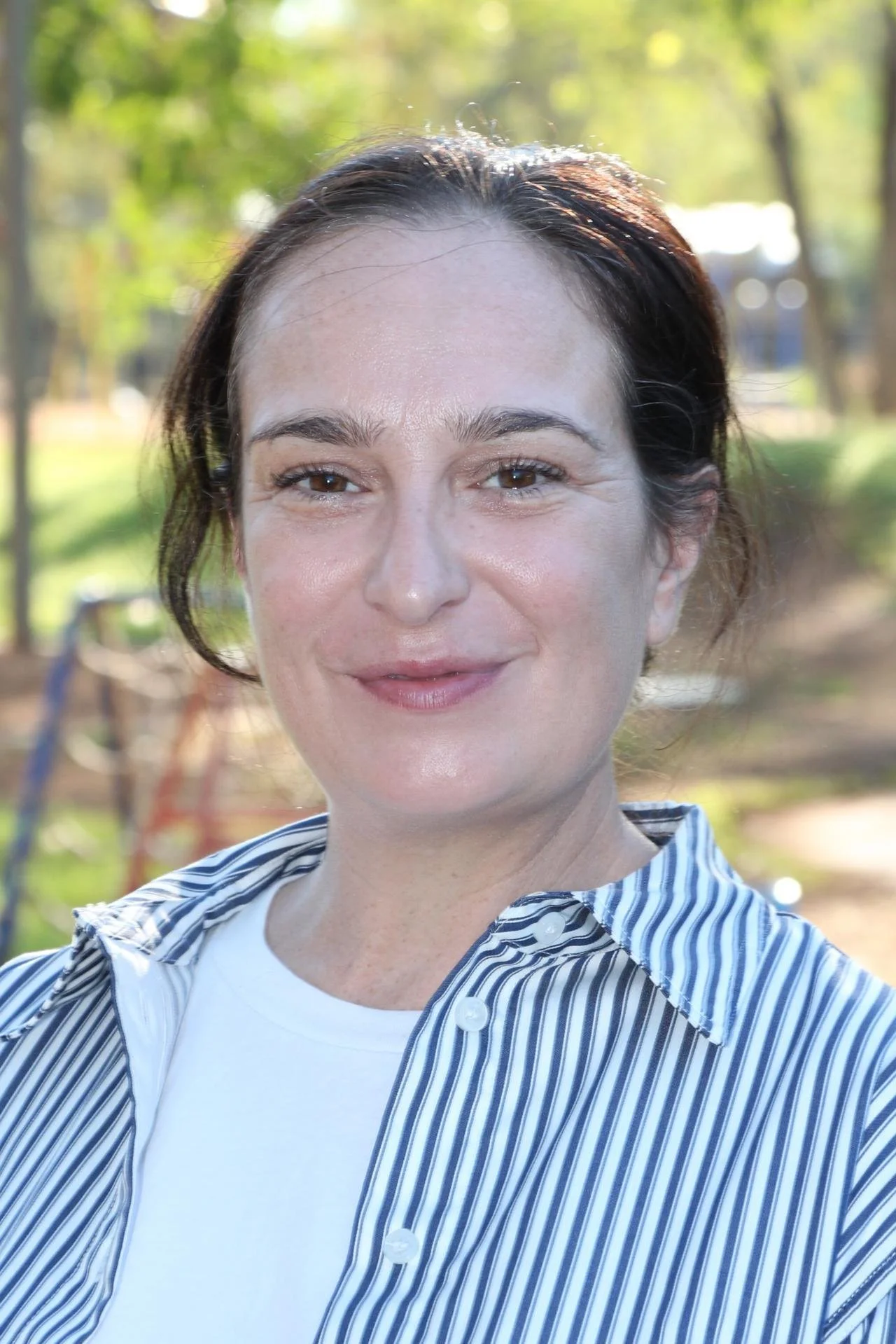 Close-up of a woman smiling outdoors in a park with green trees in the background, wearing a striped shirt and white T-shirt.