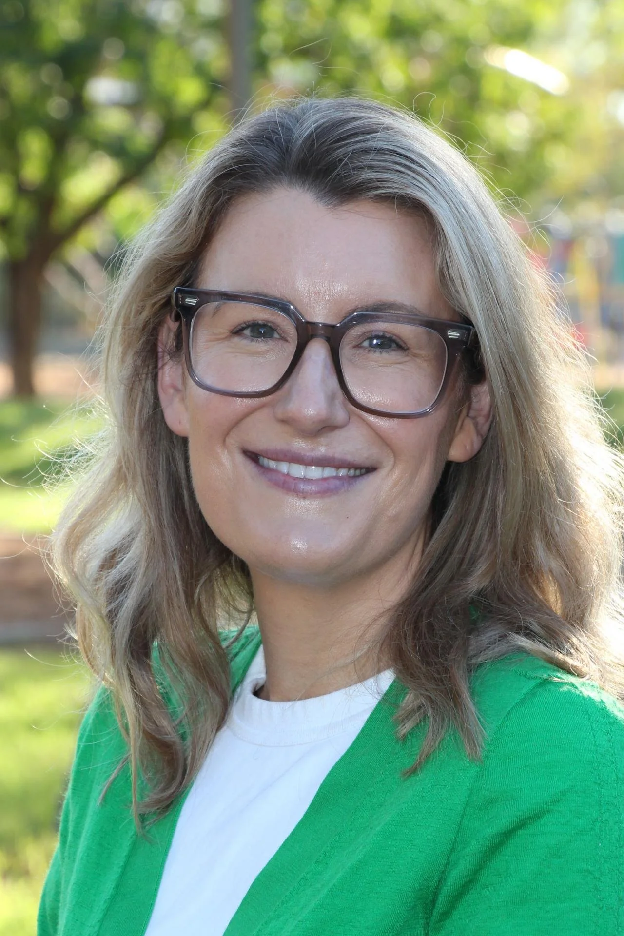 A smiling woman with blonde hair and glasses outdoors, wearing a green jacket and a white shirt, with blurred trees in the background.