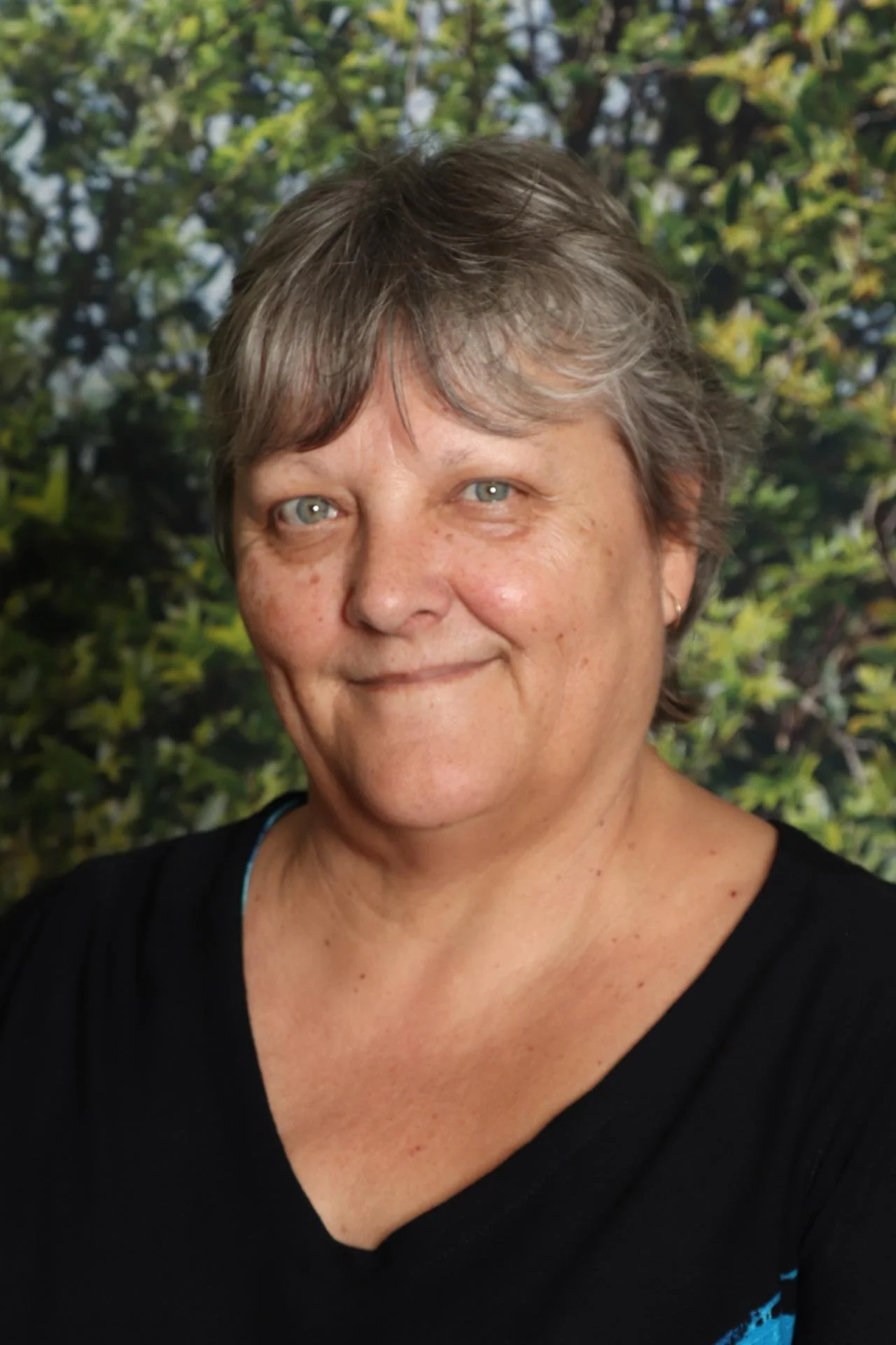A middle-aged woman with short gray hair and blue eyes smiling in front of a leafy green background.