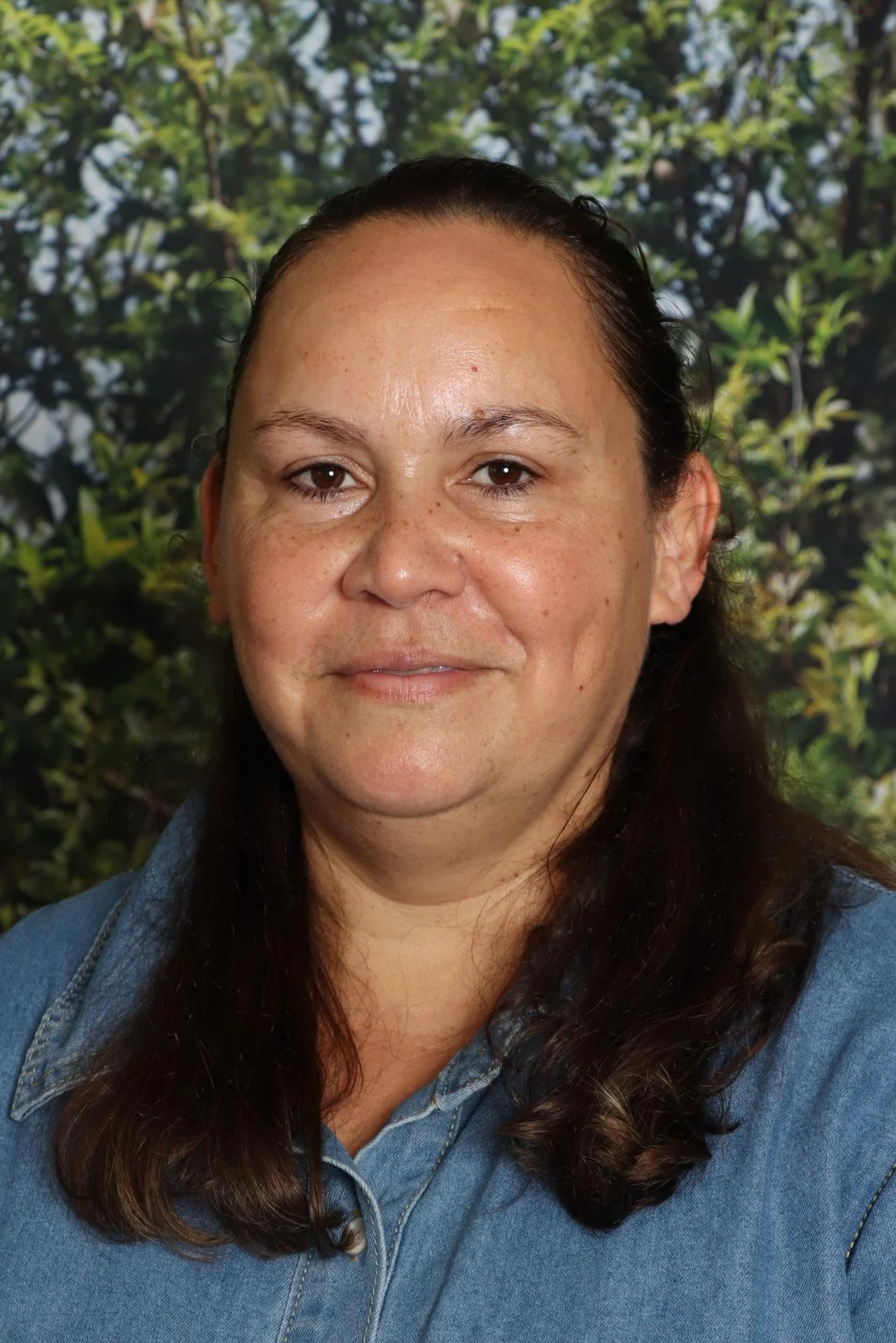 Close-up of a woman with dark brown, wavy hair wearing a blue denim shirt, smiling in front of a leafy green background.