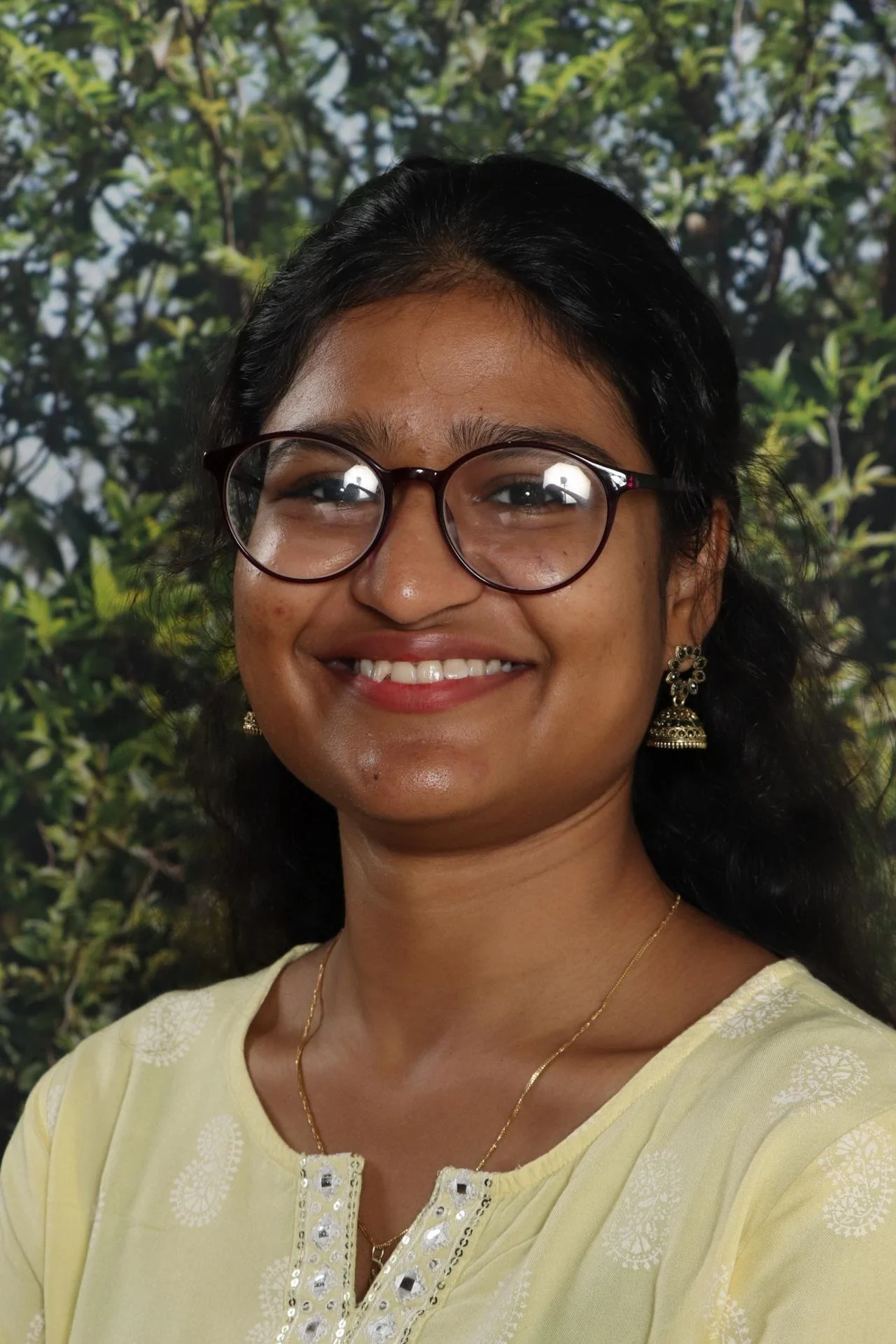 A woman with black hair and glasses is smiling in front of a green leafy background, wearing earrings and a yellow traditional outfit with white embroidery.