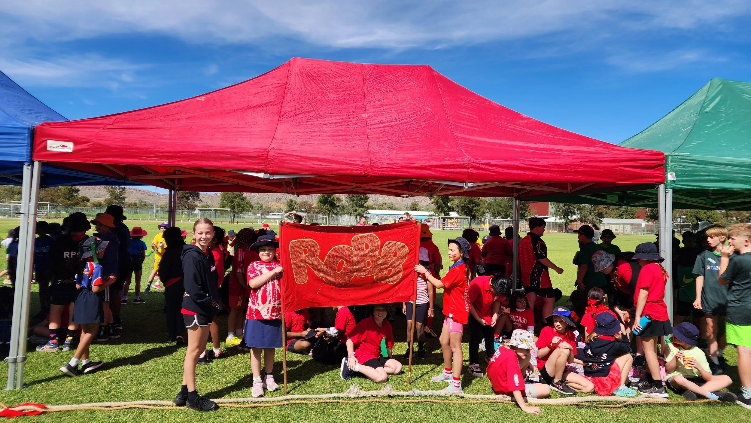 A group of children participating in an outdoor event or gathering, with some holding a red banner that says 'ROBE'. They are under colorful tents on a grassy field, wearing casual clothing and hats, with trees and clear blue sky in the background.