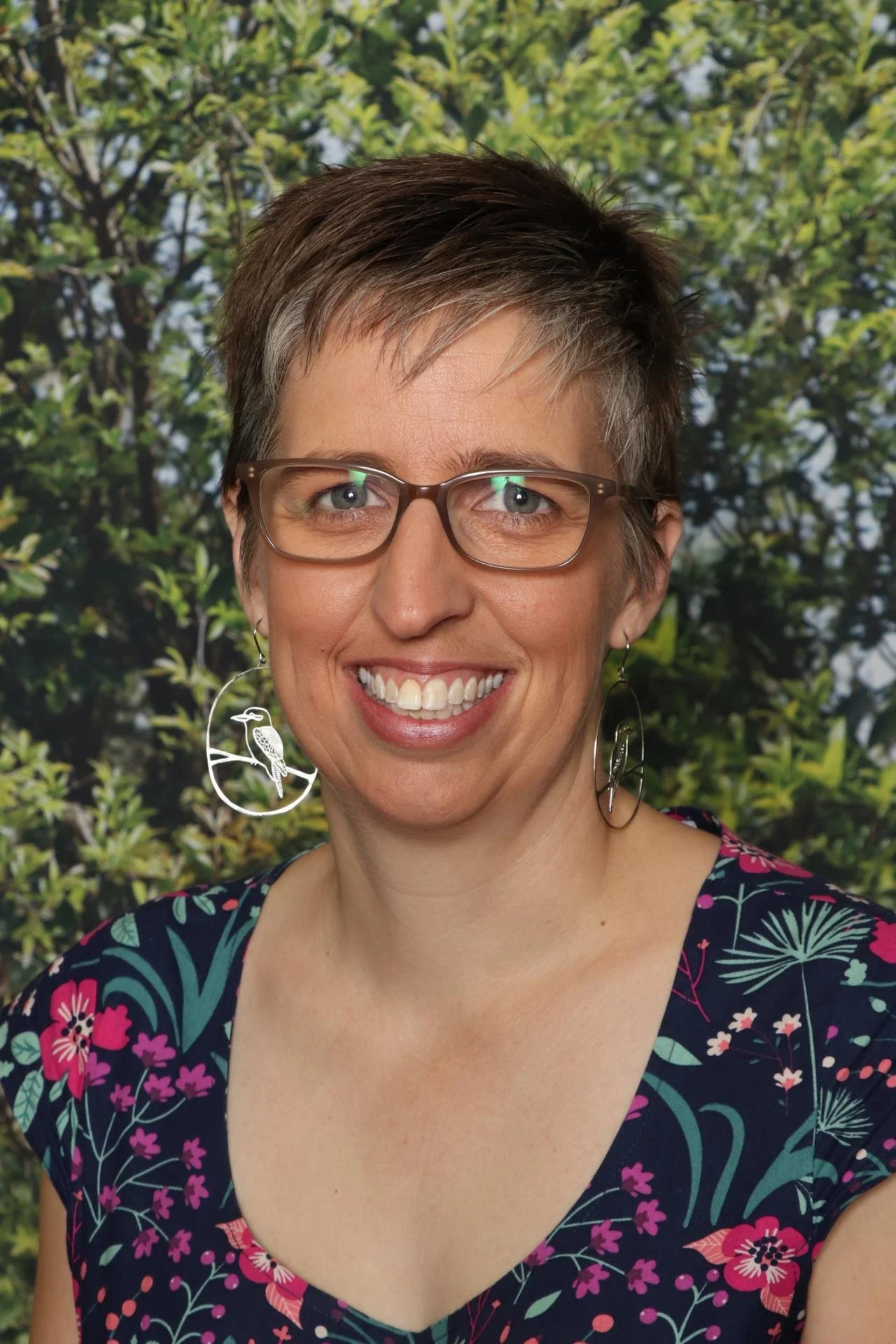 Smiling woman with short brown hair, glasses, and bird-shaped earrings, wearing a floral dress, standing outdoors with green foliage in the background.