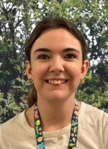 A young woman smiling outdoors with trees in the background, wearing a lanyard around her neck.
