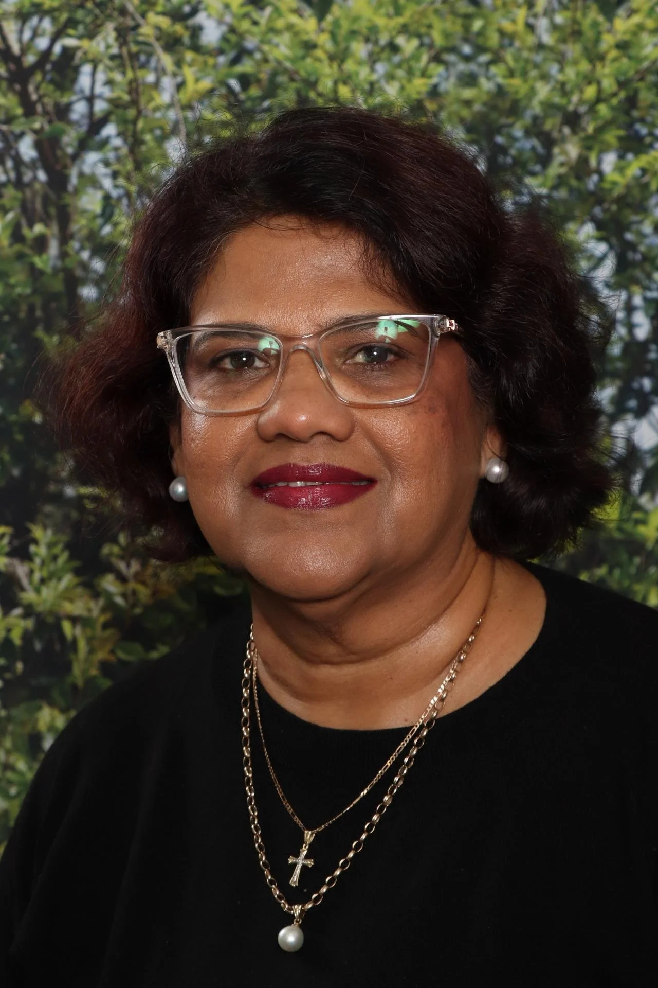 Portrait of a woman with short dark hair, glasses, red lipstick, wearing pearl earrings, layered gold necklaces, and a black top, with a green leafy background.