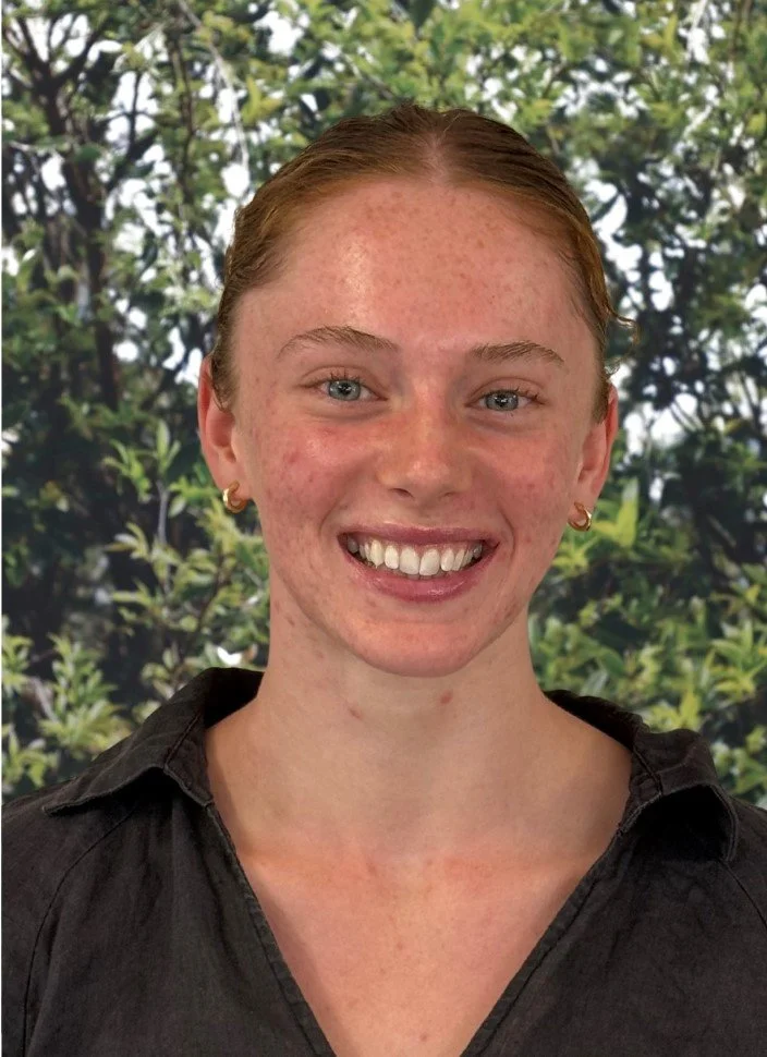 A young woman with red hair, freckles, and blue eyes smiling, wearing a black shirt and gold earrings, with a leafy green background.