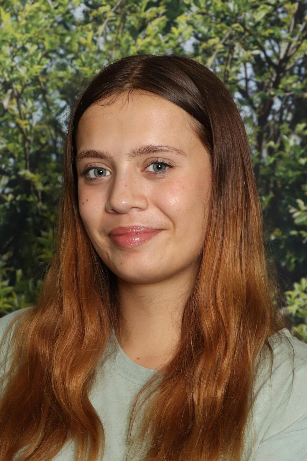 A young woman with long red hair and blue eyes smiling in front of a background of green leaves