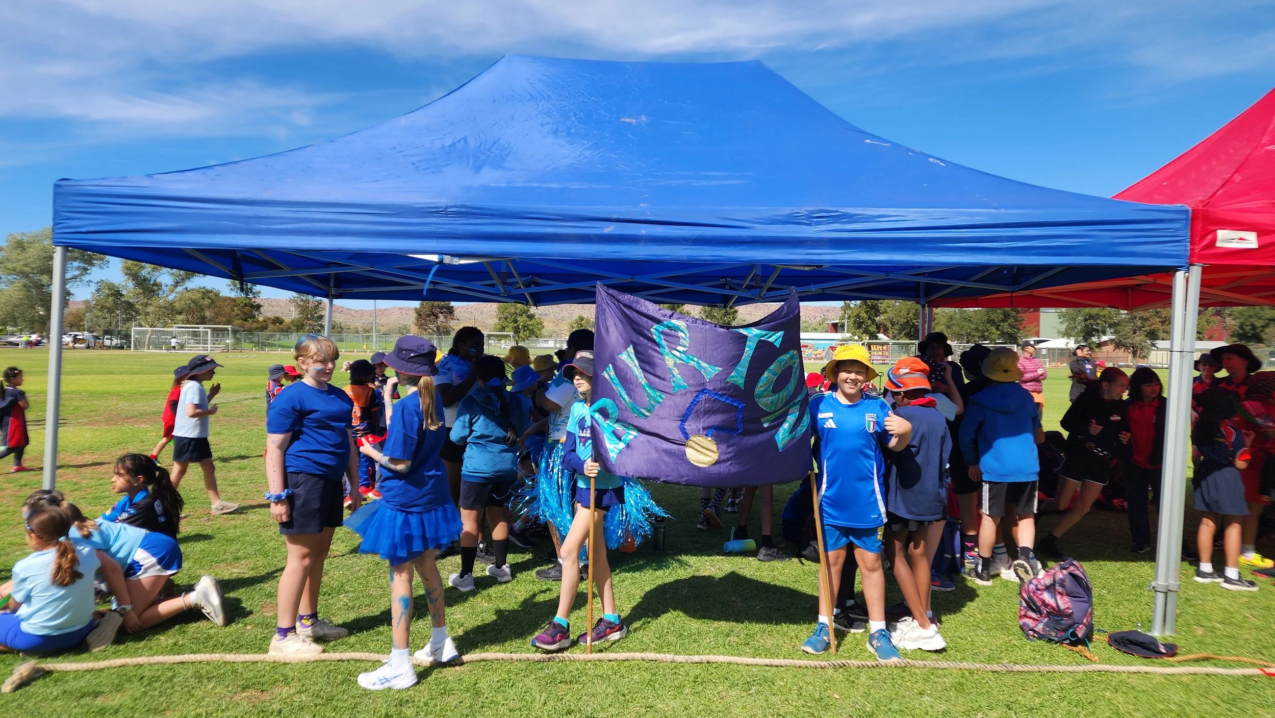 Children gathered under a blue tent on a grassy field during a sunny outdoor event, some holding a sign that says 'RURI' with colorful decorations.