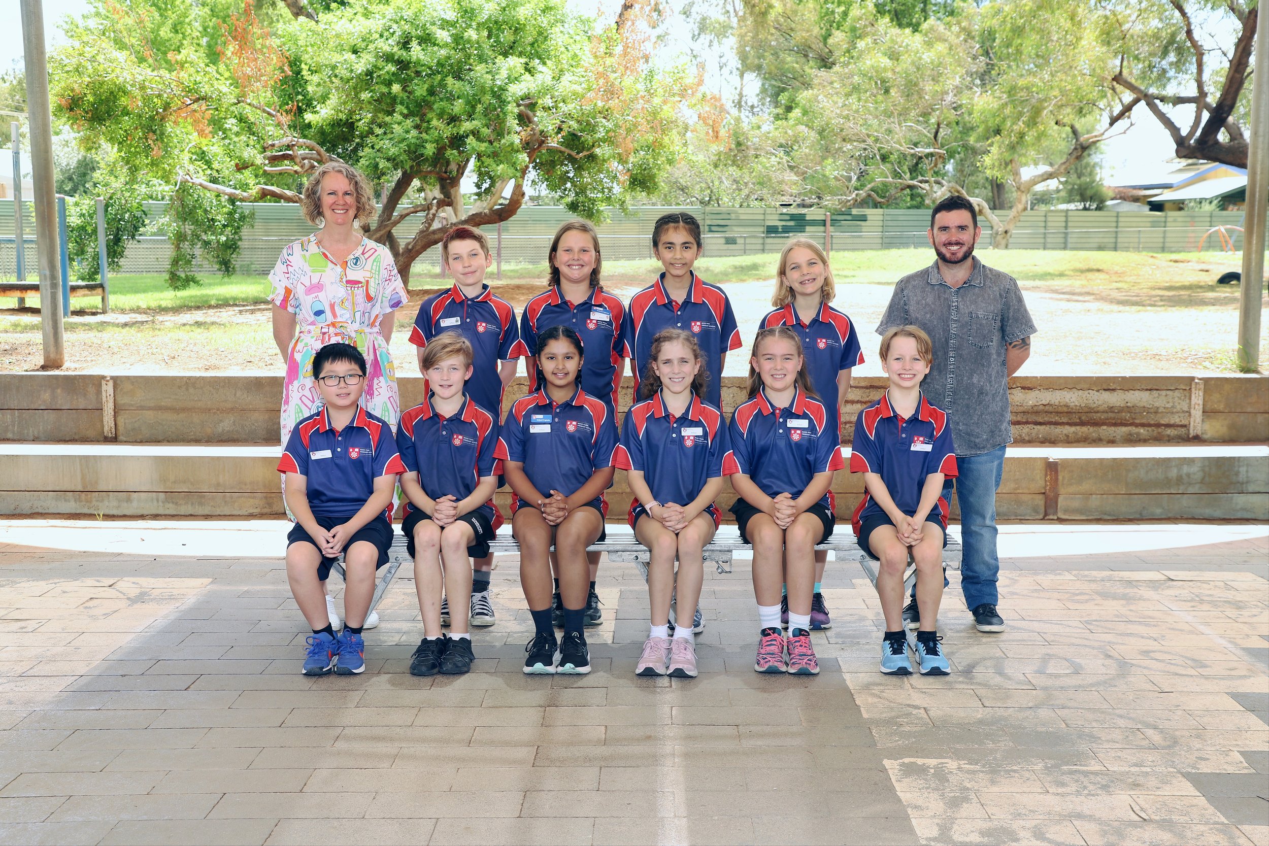 Group photo of eight children and two adults outdoors on brick patio, with trees and a metal fence in the background. The children are in matching red and blue sports uniforms, and one adult woman is wearing a colorful dress, while the man is in a gray shirt. All are smiling and posing for the picture.