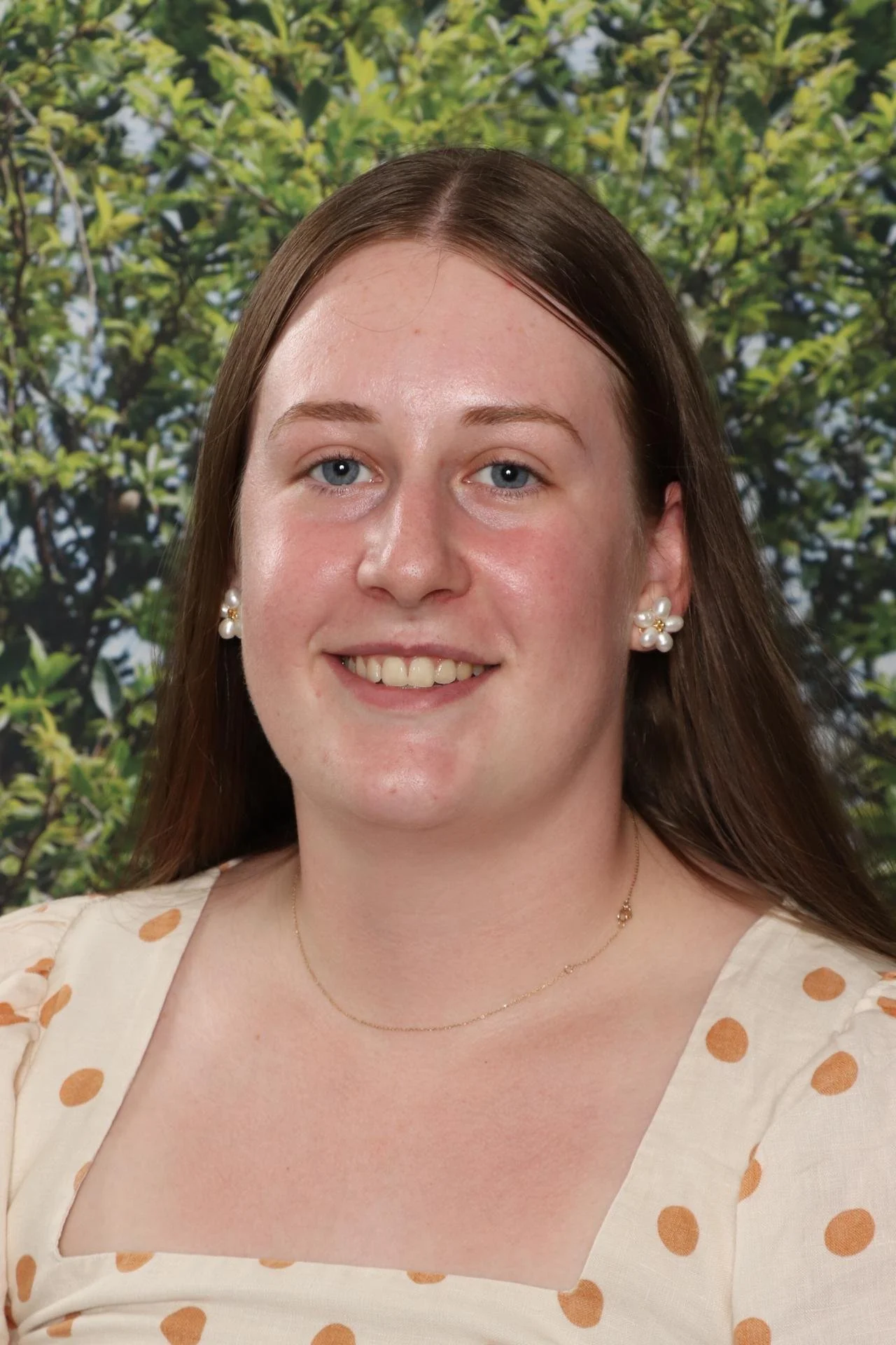 A woman with brown hair, blue eyes, and earrings smiling in front of a leafy background.