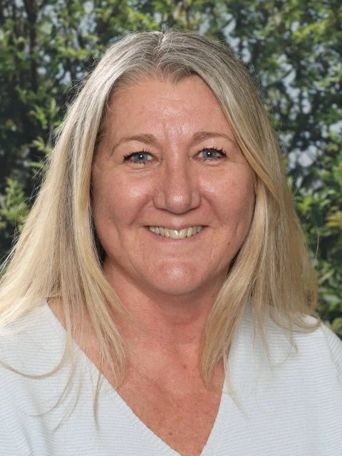 A smiling middle-aged woman with long blonde hair, wearing a white top, standing outdoors with blurred green trees in the background.