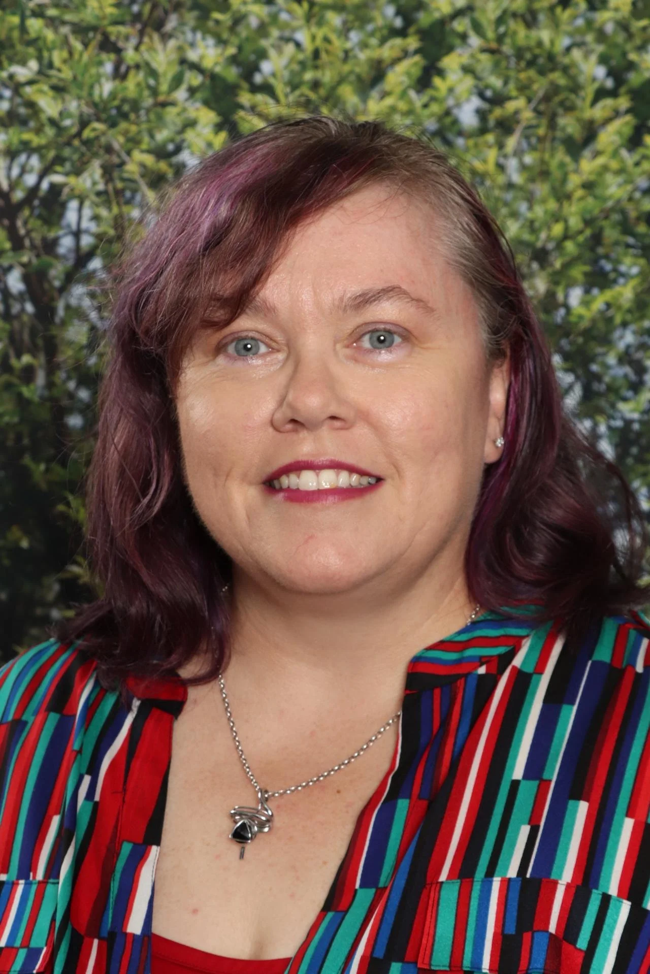 A woman with shoulder-length dark red hair and blue eyes, wearing a colorful striped shirt and a necklace with a key and butterfly pendant, standing outdoors with green foliage in the background.