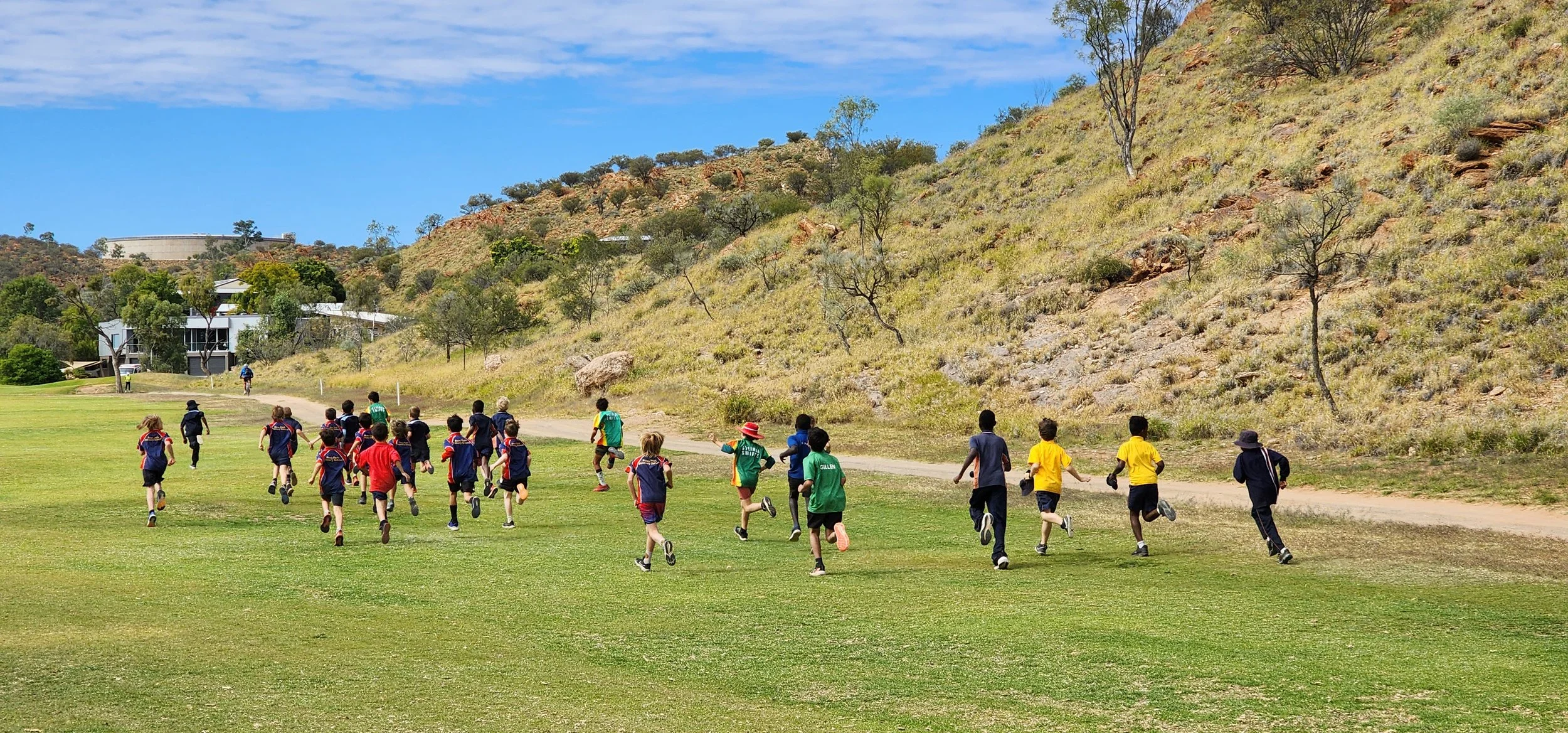 Children wearing school uniforms running on a grassy field with a dirt pathway, surrounded by rocky hills and sparse trees under a blue sky.