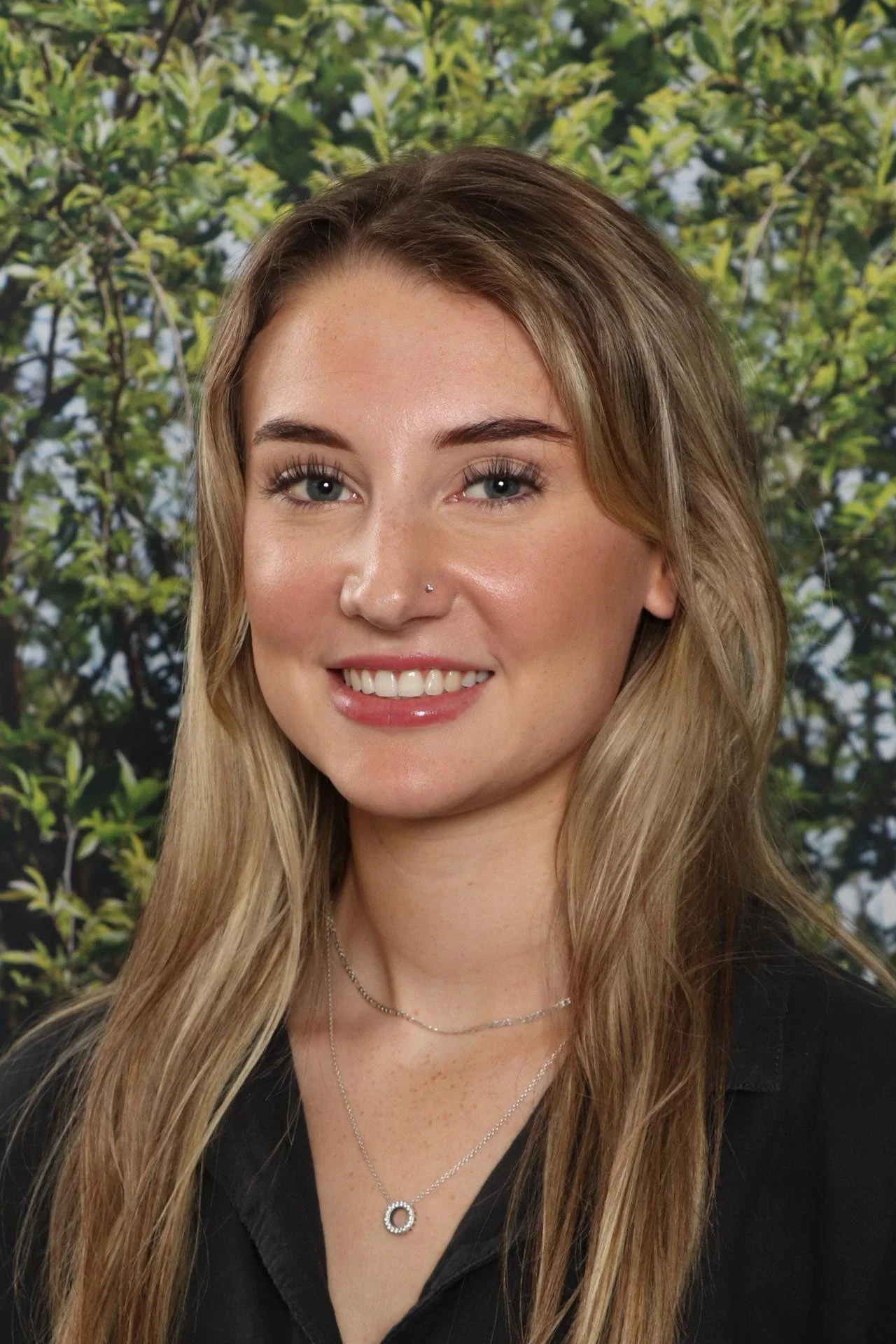 Close-up portrait of a young woman with long, wavy blonde hair, blue eyes, and a nose piercing, smiling in front of a background of greenery.