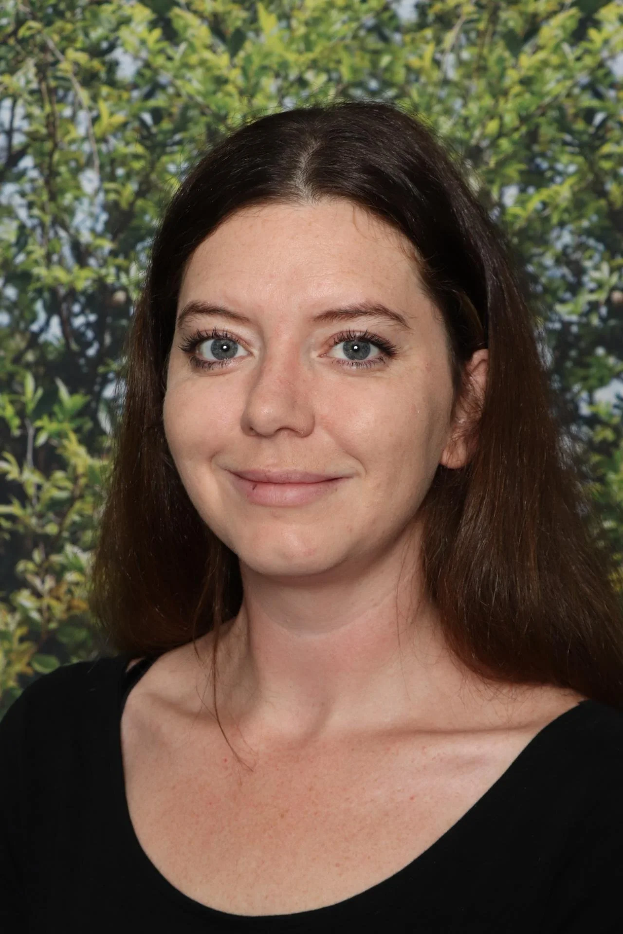 A woman with long dark hair and blue eyes, wearing a black top, standing in front of a background of green leaves.