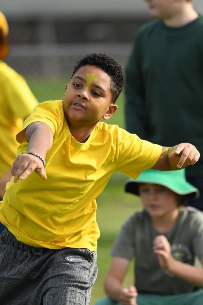 Young boy in a yellow shirt running outdoors, with face paint and a bracelet, participating in a race or outdoor activity. Other children are in the background.