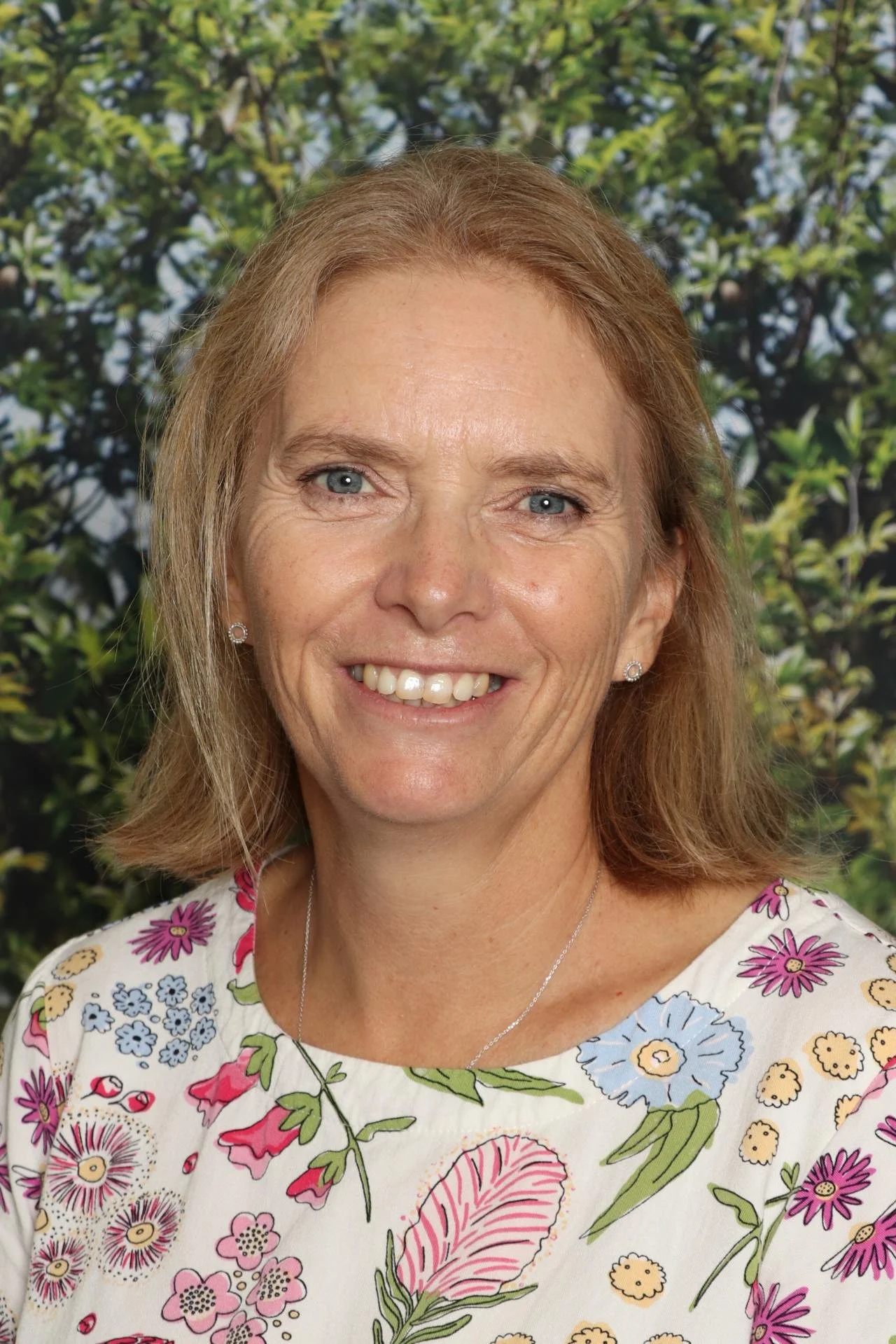 A smiling woman with light brown hair and blue eyes is wearing a floral patterned shirt with pink, purple, and green flowers. The background consists of green foliage.