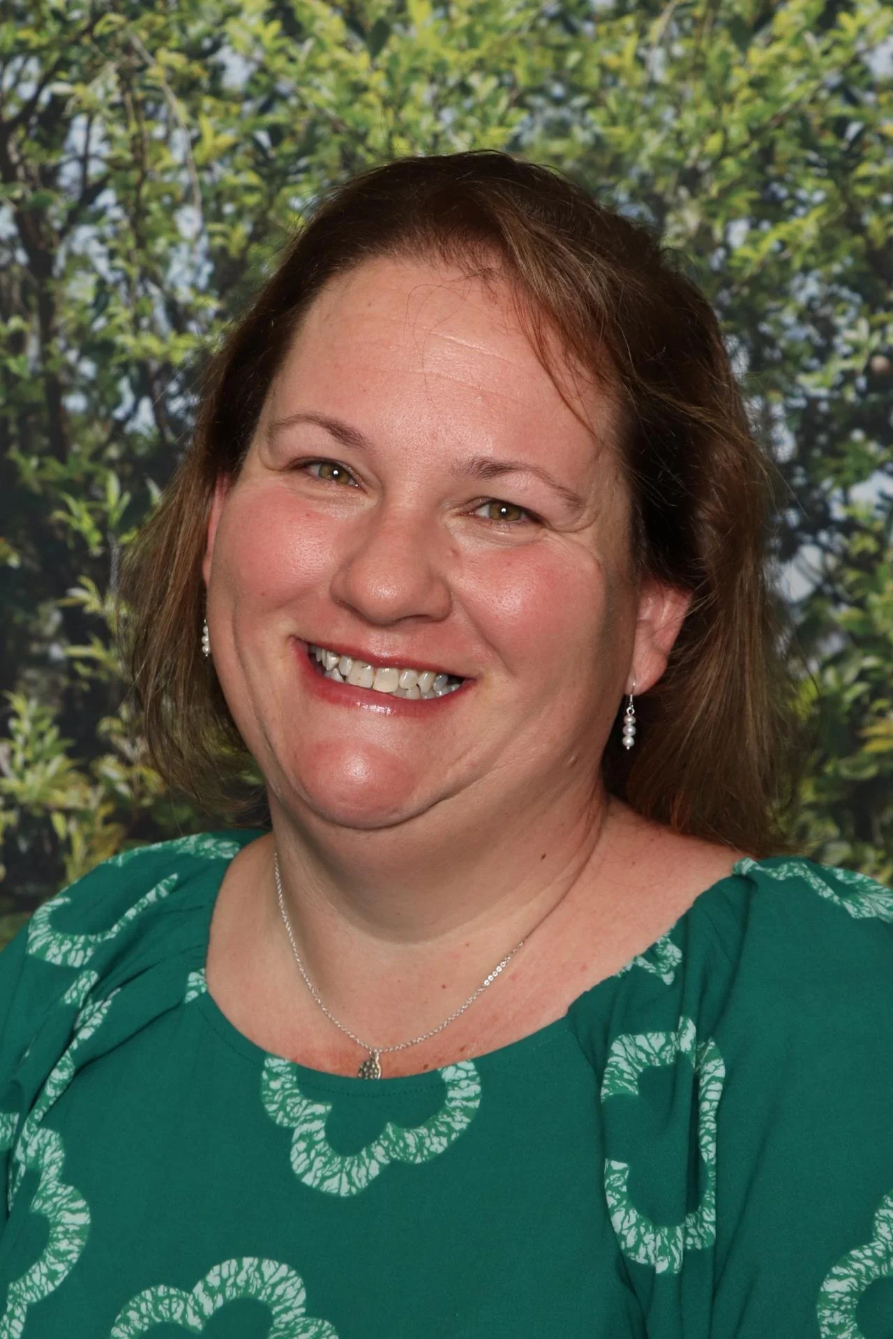 A smiling woman with shoulder-length brown hair, wearing a teal dress with a white floral pattern, pearl earrings, and a silver necklace, standing in front of a background with green trees.