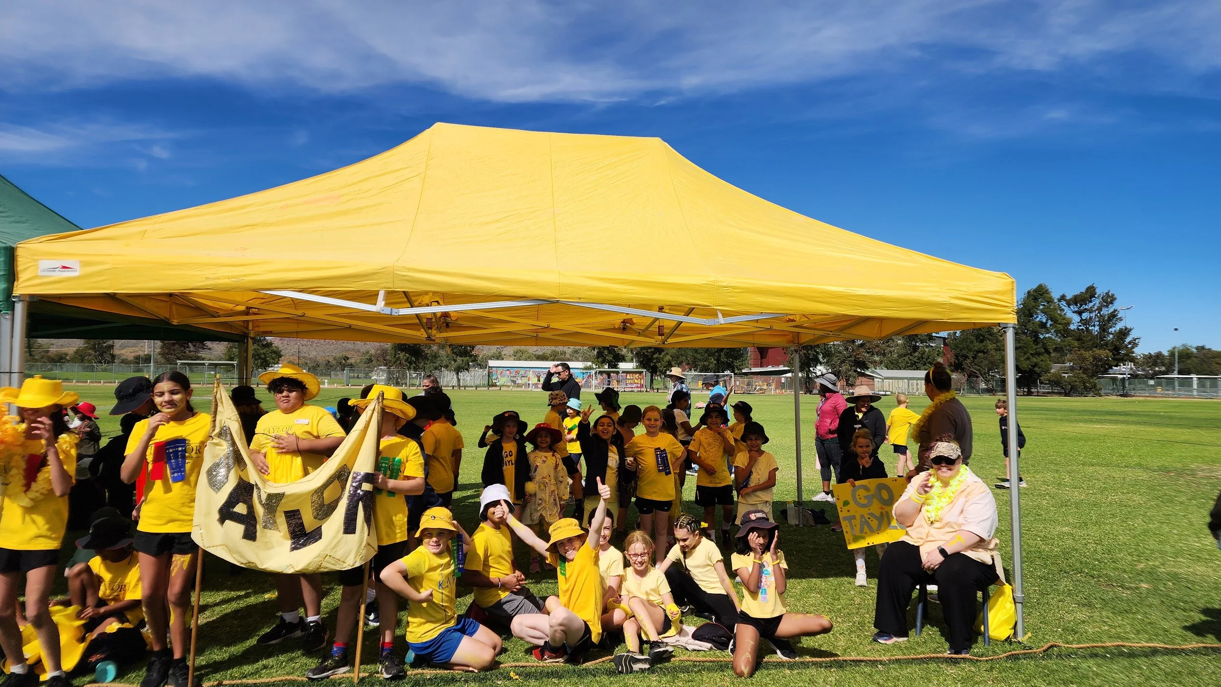 Group of children and adults gathered under a yellow tent outdoors, some children wearing yellow shirts and hats, holding signs and prizes, on a grassy field with trees and a blue sky in the background.