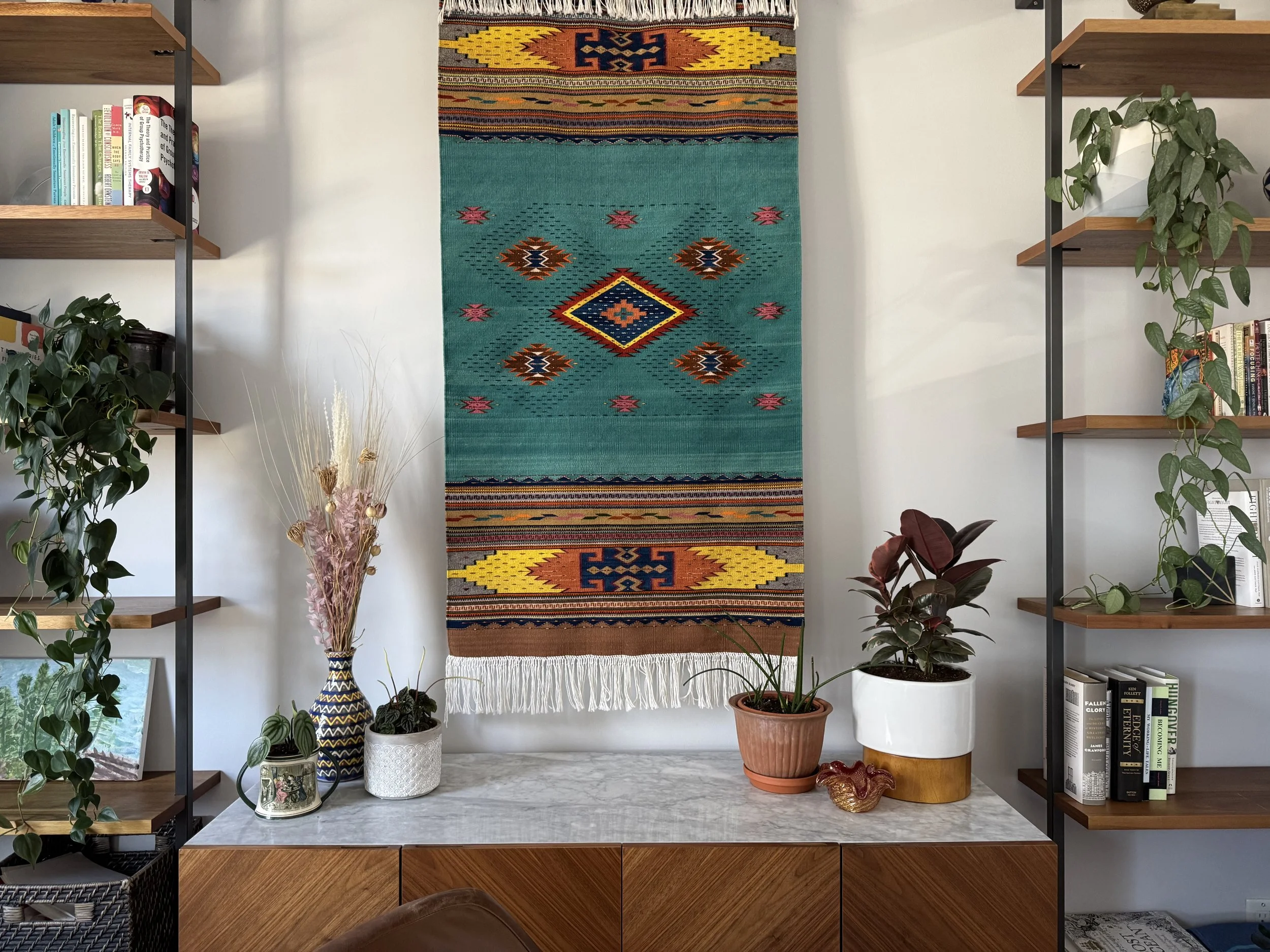 An home office with plants on a marble drawer, with a ornate rug hanging above.
