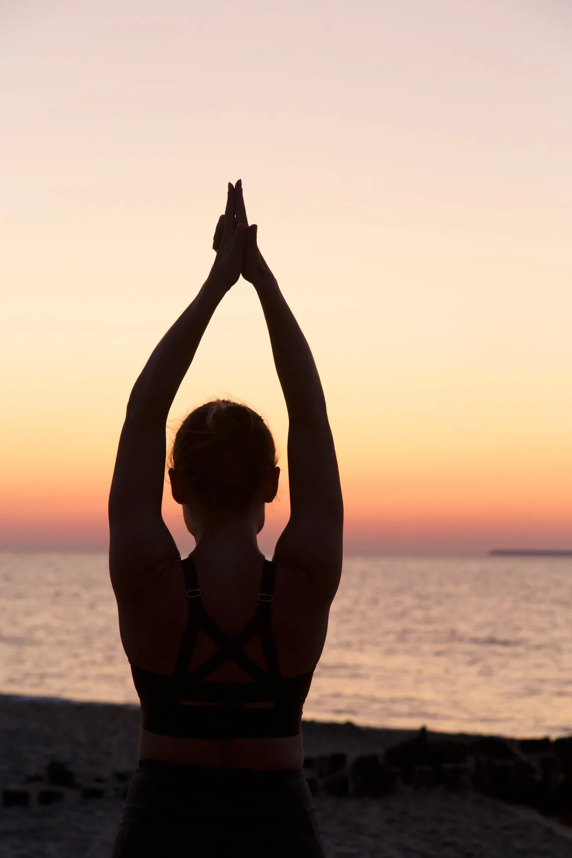 Silhouette of a woman practicing yoga on the beach during sunset, with her hands in prayer position above her head.