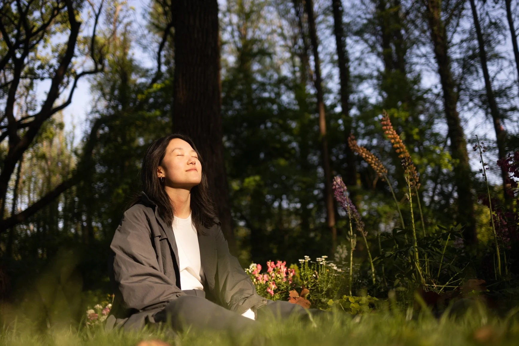 A woman sitting cross-legged outdoors in a forest, eyes closed, enjoying sunlight.