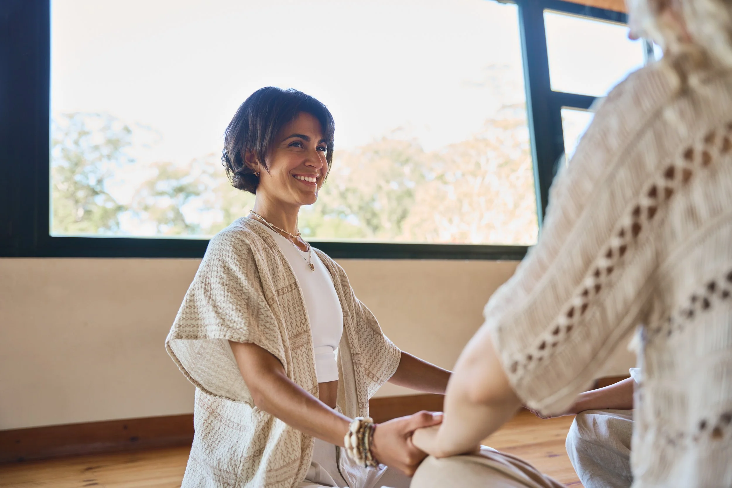 A woman smiling happily while sitting cross-legged on the floor, holding hands with another person whose face is not visible. They are in a room with large windows and a view of trees outside.