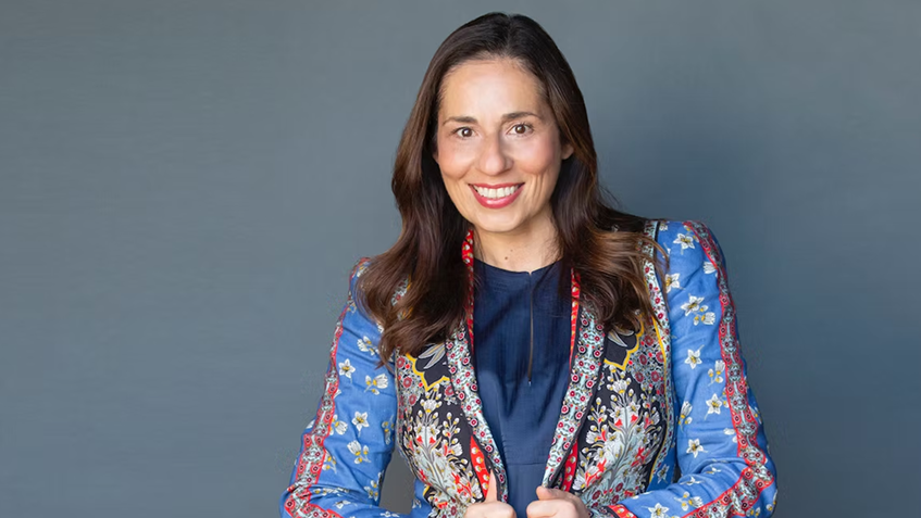 A woman with long brown hair wearing a colorful, patterned jacket over a navy top, smiling against a gray background.