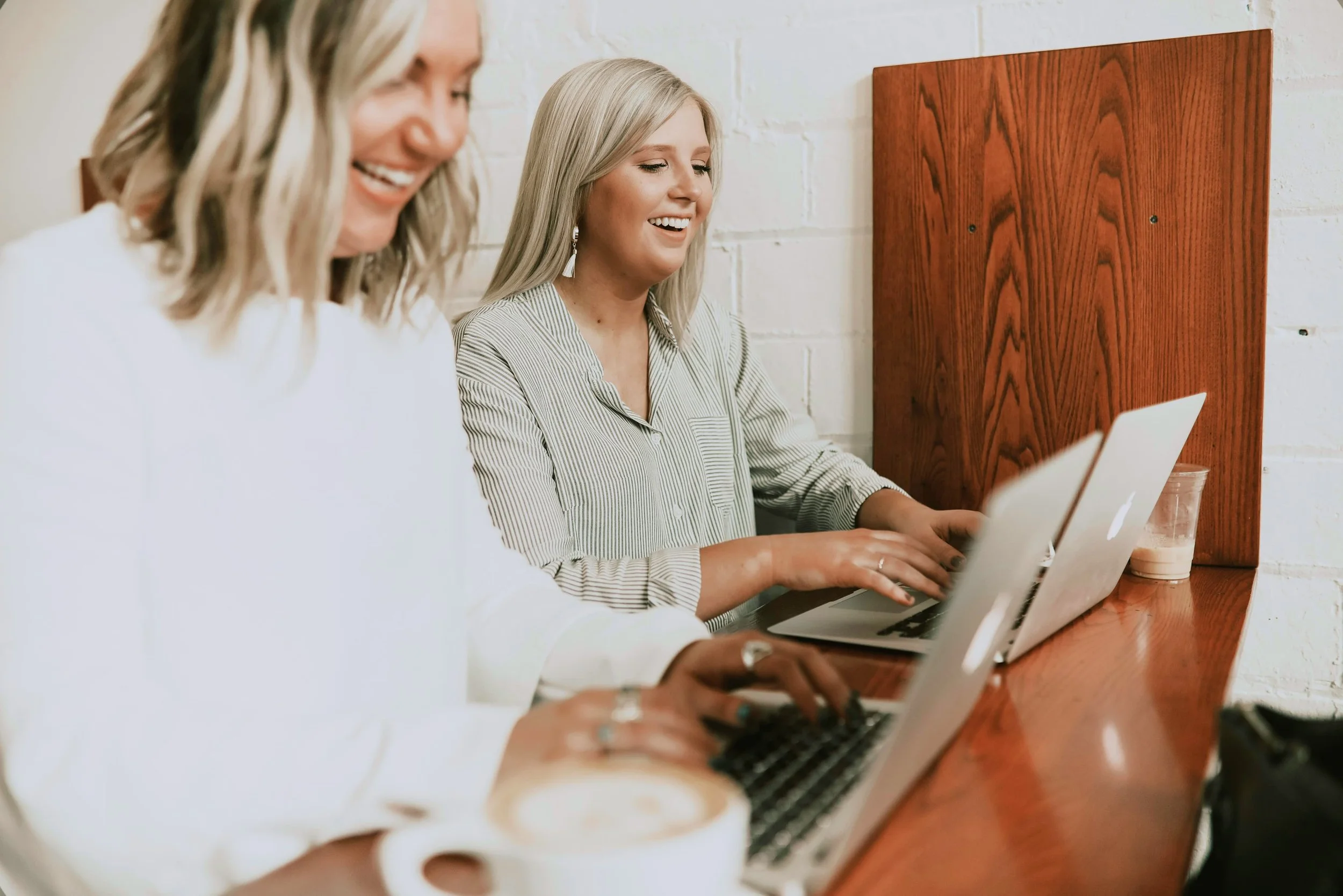 Two women sitting at a wooden table working on laptops, smiling, with coffee cups nearby, against a white brick wall.