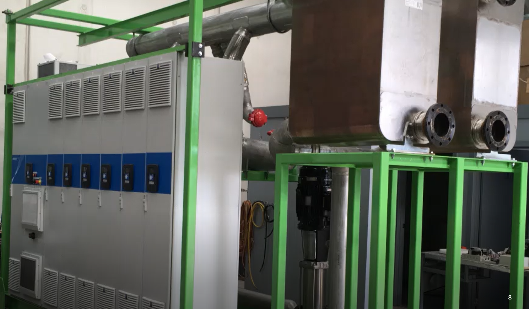 Industrial equipment including a large gray machine with vents, pipes, valves, and a green metal framework surrounding parts of the machinery.