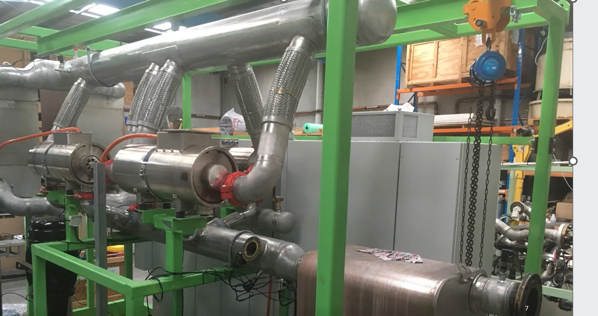 Industrial piping system with large stainless steel pipes, valves, and metal fittings, supported by a green metal framework in a workshop or plant setting.