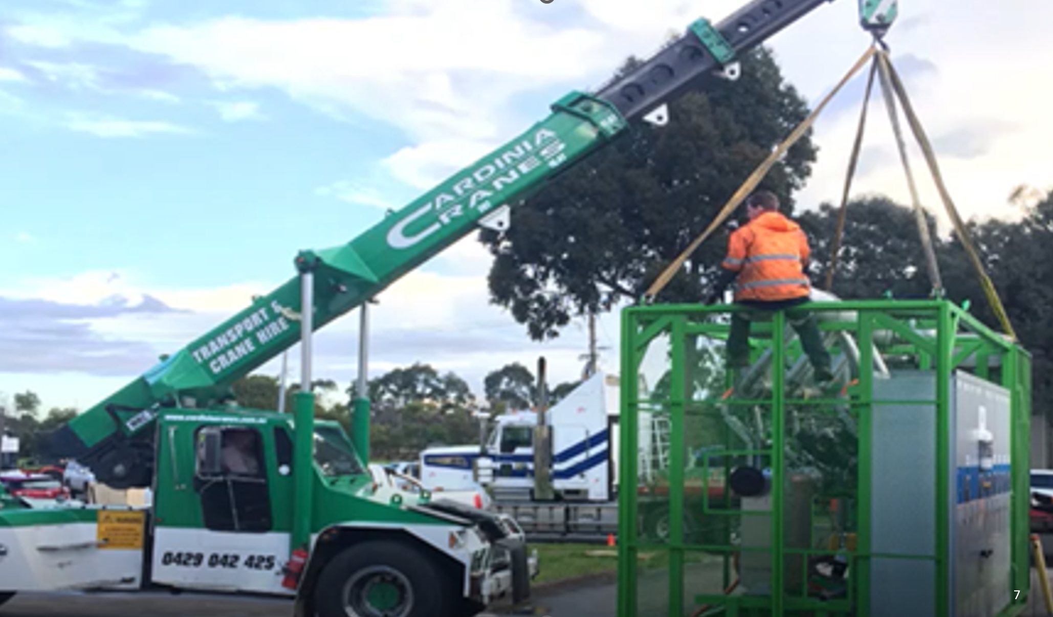 A worker operating a green cherry picker lift with the words "Cardinal Crane" on its arm, lifting a large utility pole or crane component in a parking lot area with various trucks and vehicles in the background.