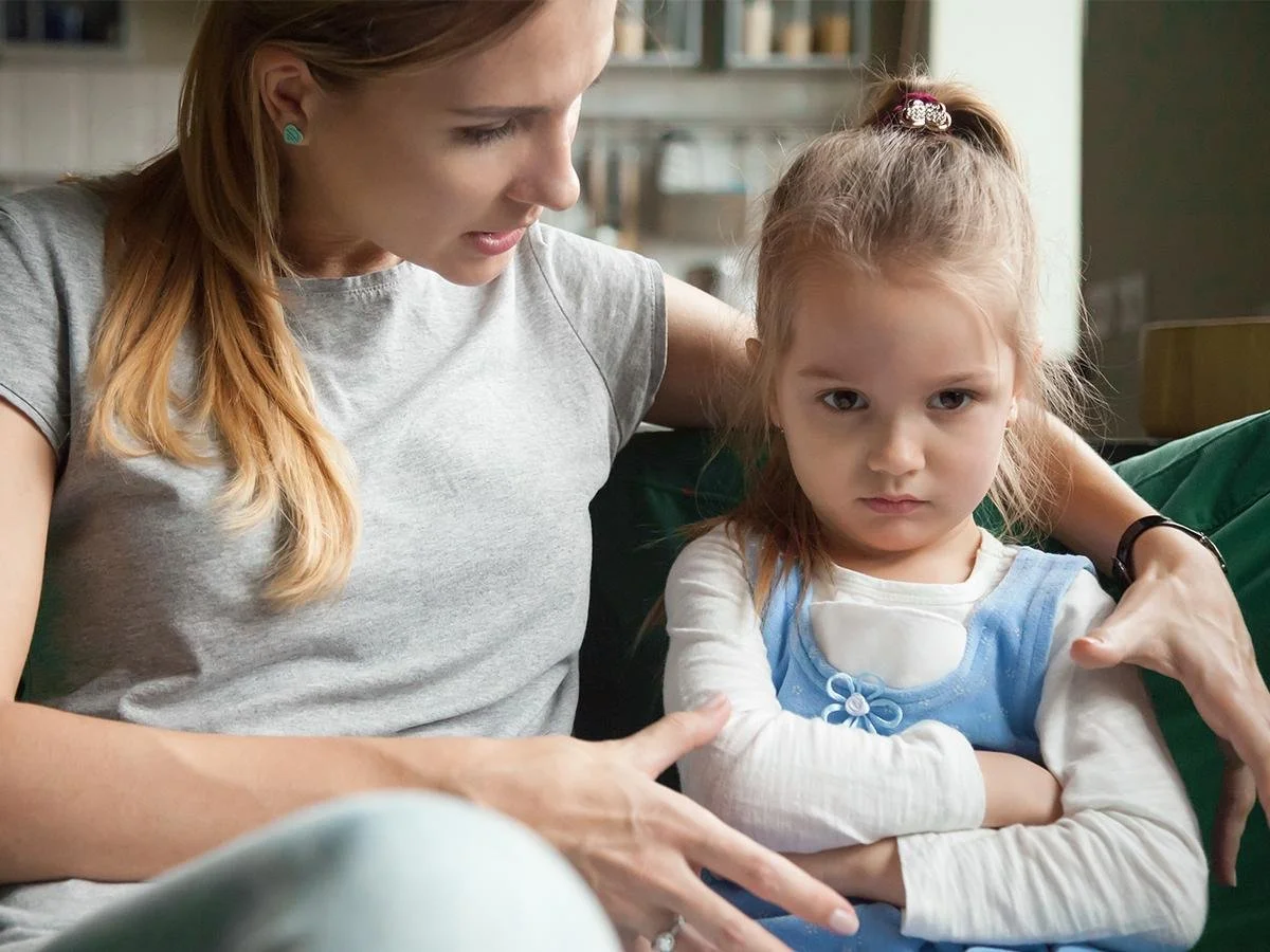 A woman appears to be scolding or explaining something to a young girl with crossed arms and a serious expression, sitting on a green sofa in a cozy room.