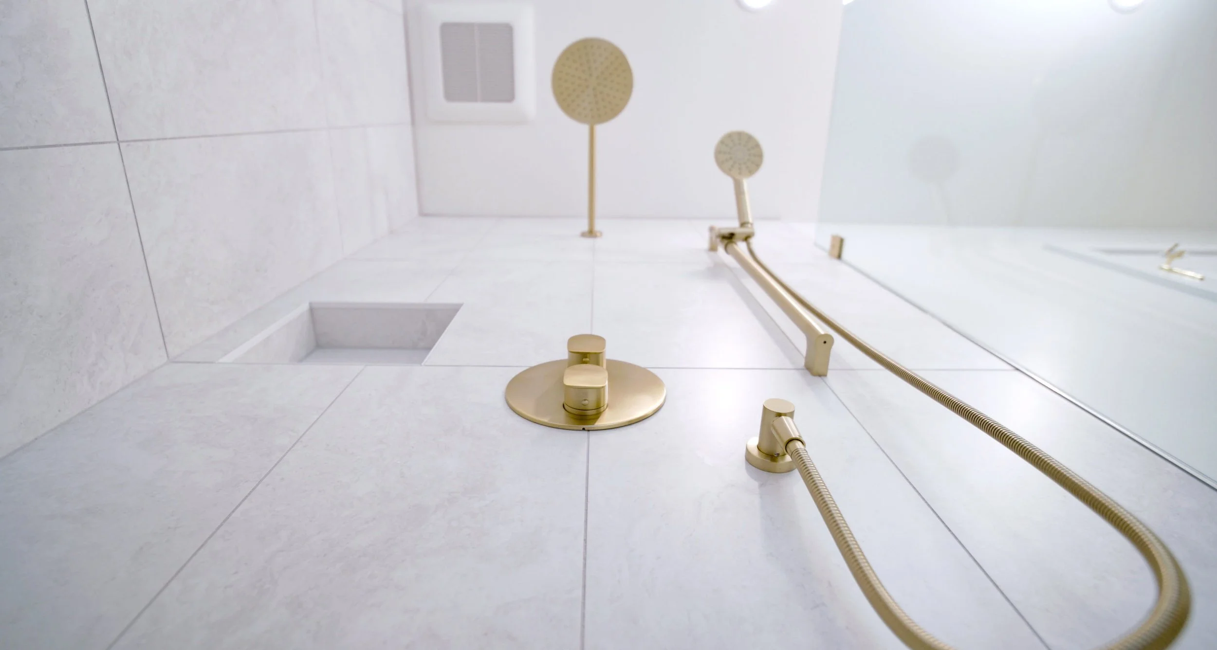 A modern shower area with beige fixtures, handheld showerheads, a wall-mounted showerhead, and a ceiling vent, all surrounded by white tiles.