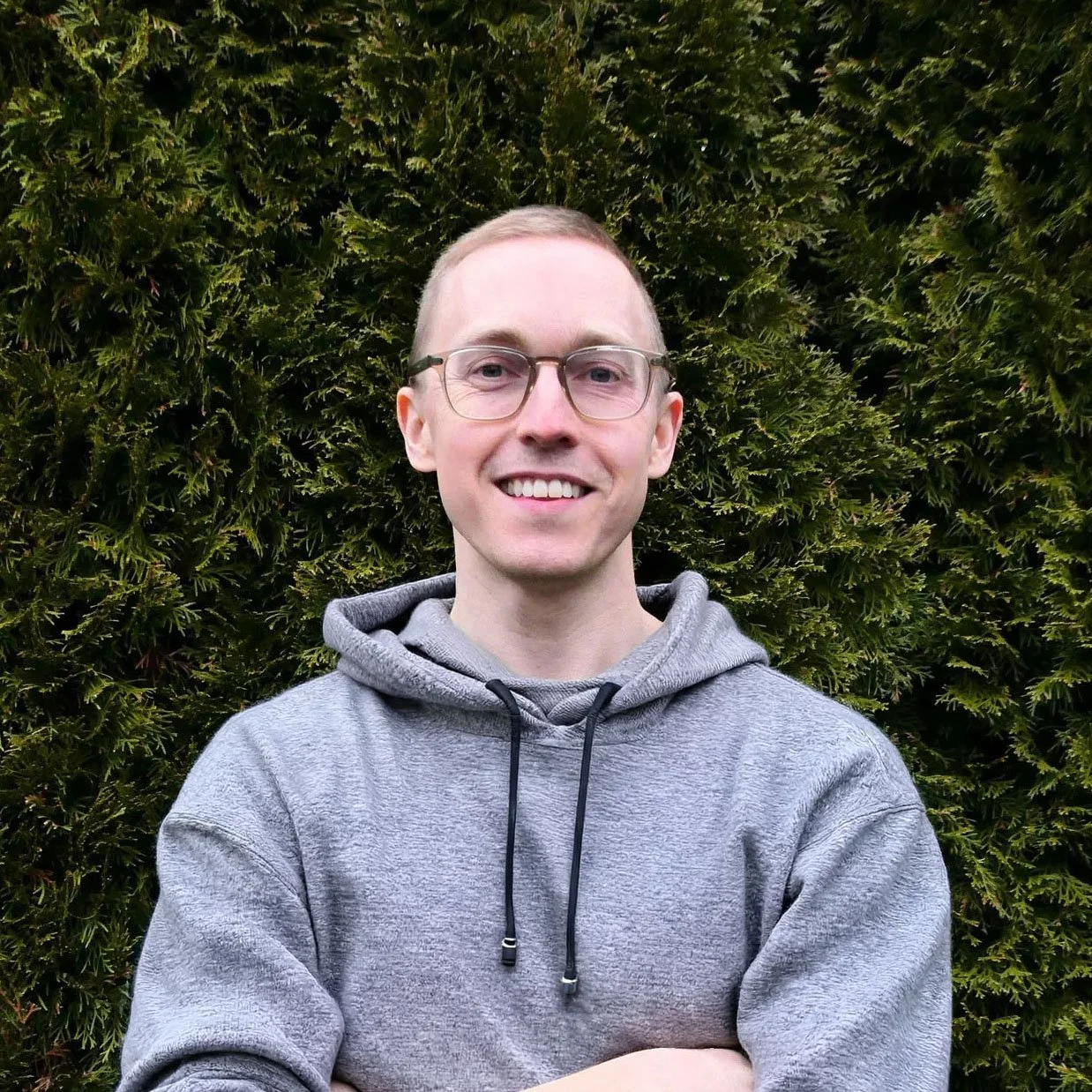 Leeve Comeau, owner of Gneiss Contracting. Leeve has short blonde hair, wearing glasses and a grey hoody, standing in front of a green leafy background