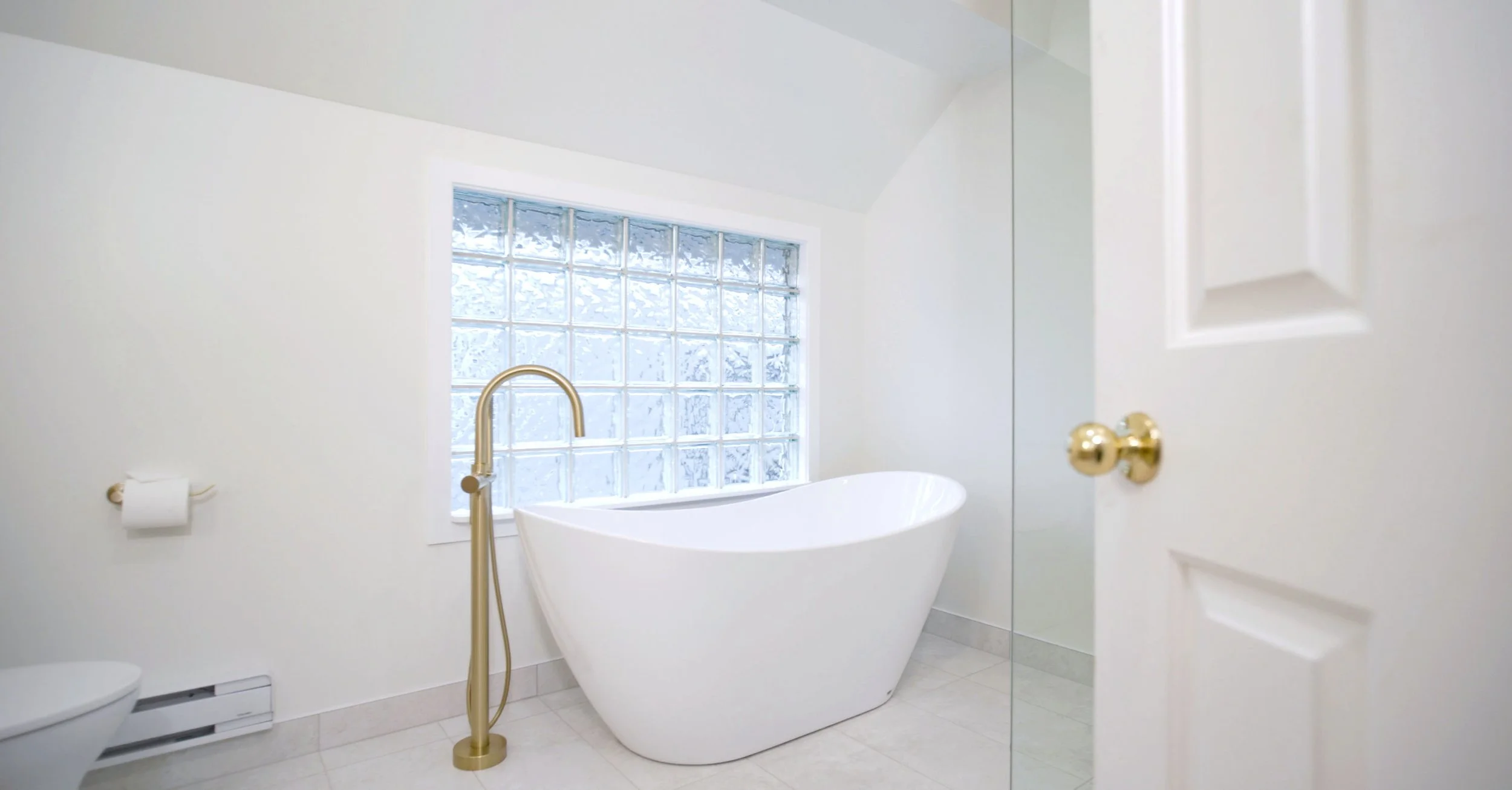 White freestanding bathtub with gold floor-mounted faucet in a bright bathroom with glass block window, white walls, and beige tile floor