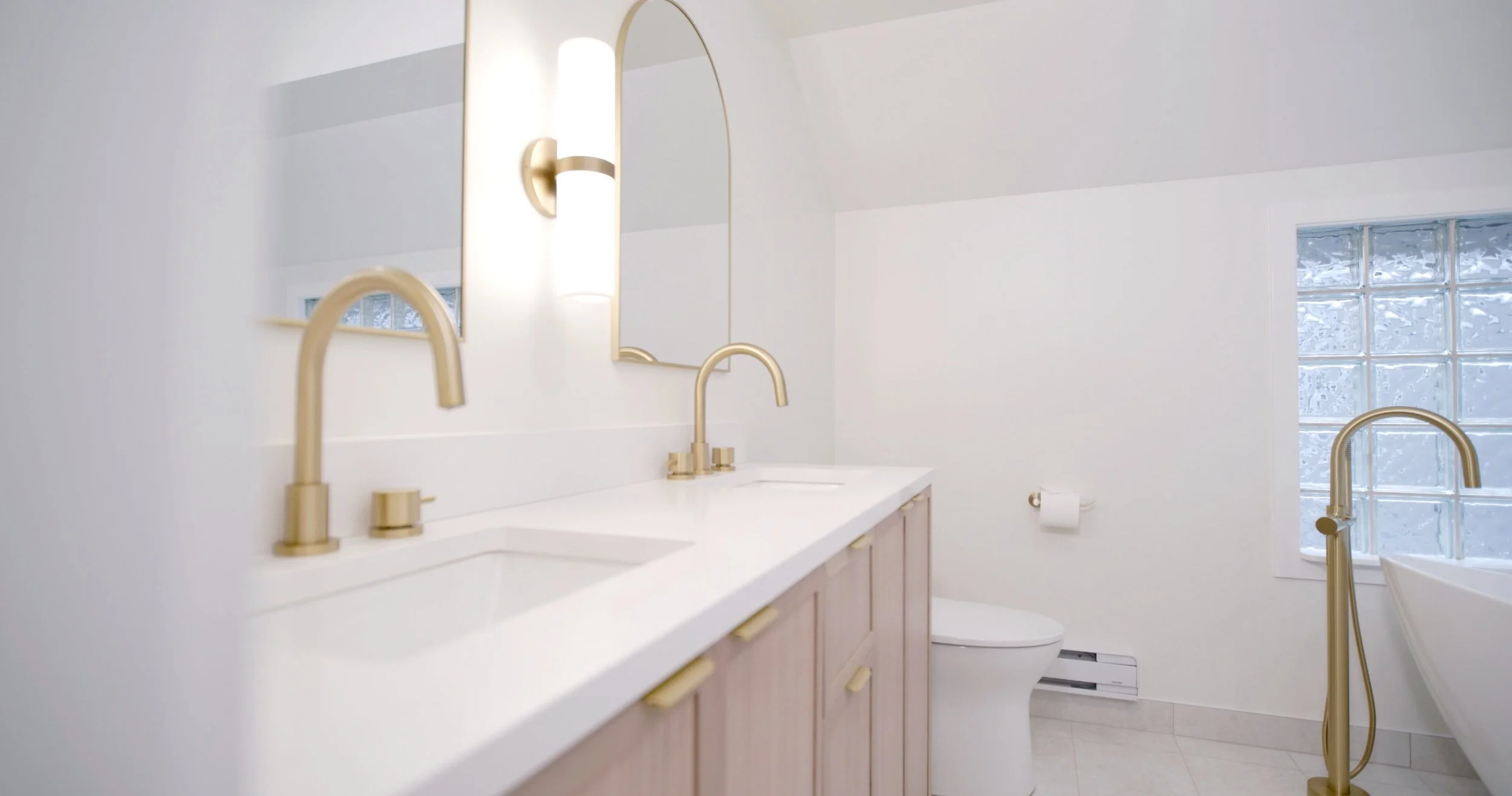 Modern bathroom with white walls, gold fixtures, dual sinks, large mirror, window with frosted glass blocks, and a toilet.