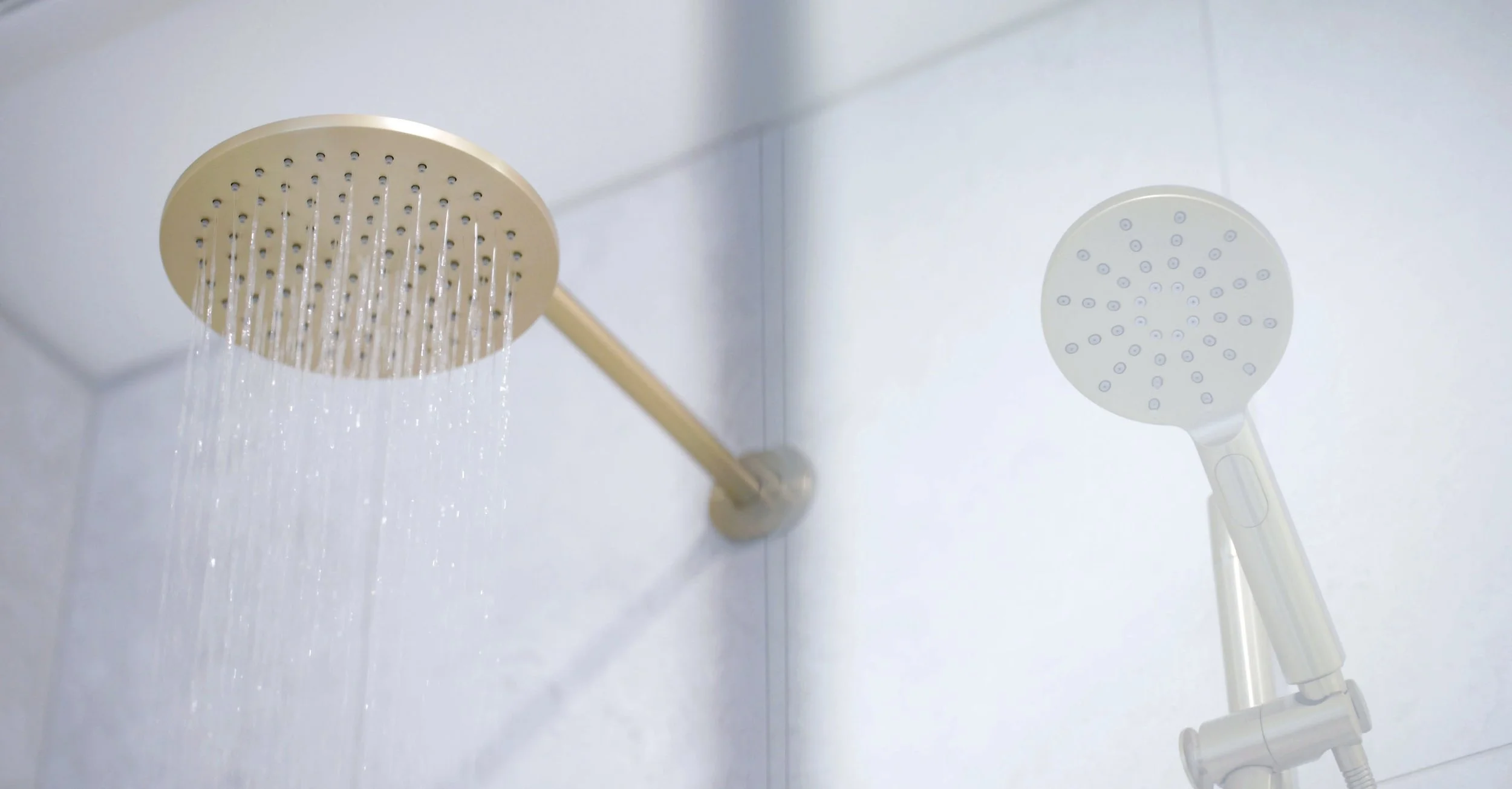 A handheld shower head and a ceiling-mounted shower head in a bathroom with white tiles, with water flowing from both.