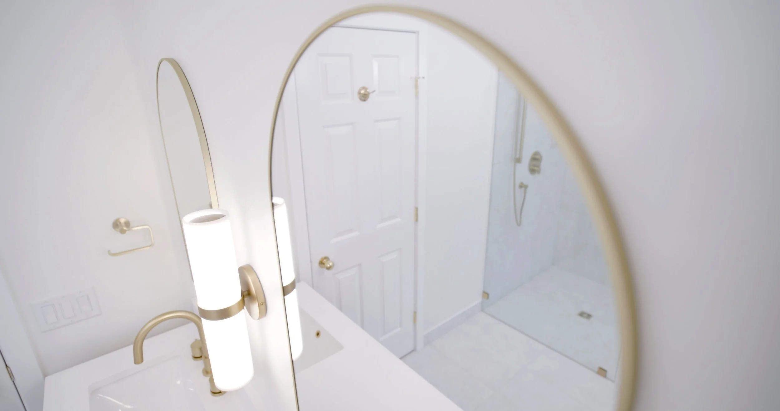 A bathroom sink and mirror with beige fixtures, a door with a doorknob, and a light switch on the wall.
