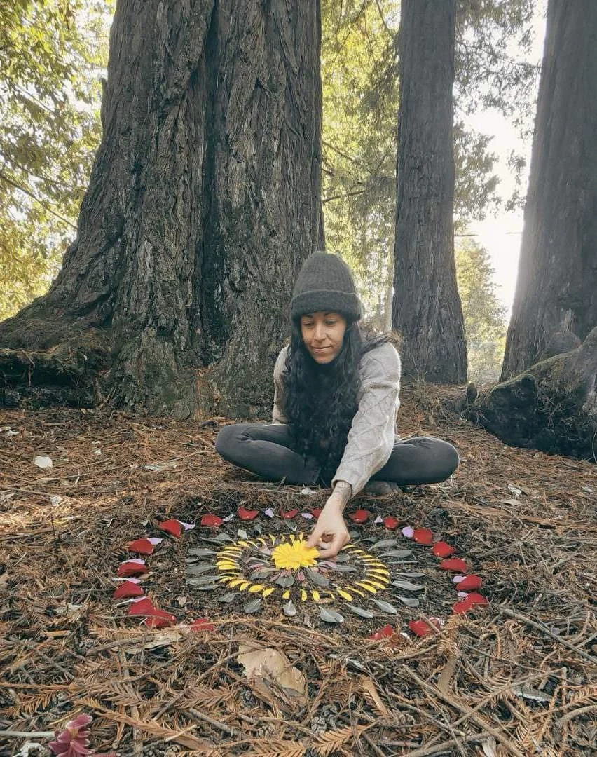 Une femme assise dans la forêt, créant un mandala avec des feuilles, des pétales de fleurs et un centre en forme de soleil en utilisant des éléments naturels sur le sol forestier.