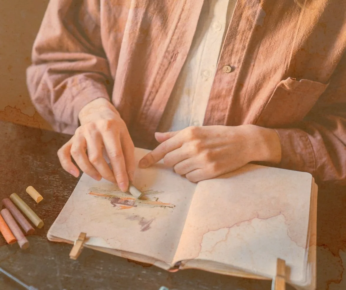 Une personne coloriant dans un carnet avec des craies de différentes couleurs posées à côté.