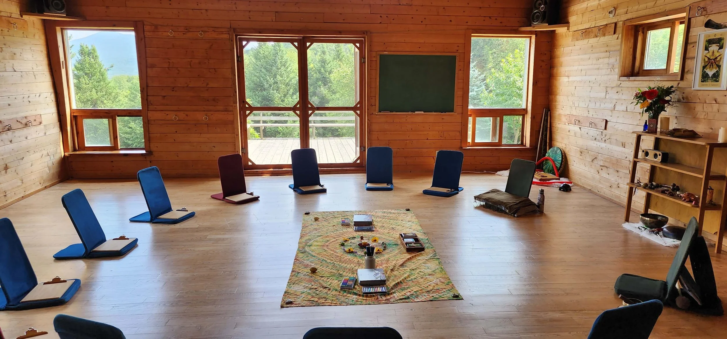 A spacious, wooden yoga or meditation room with large windows showing greenery outside. Chairs are arranged in a circle, and a colorful cloth with books, candles, and art supplies is in the center. There is a chalkboard, a bookshelf with decorative items, and a small balcony visible through the central glass door.