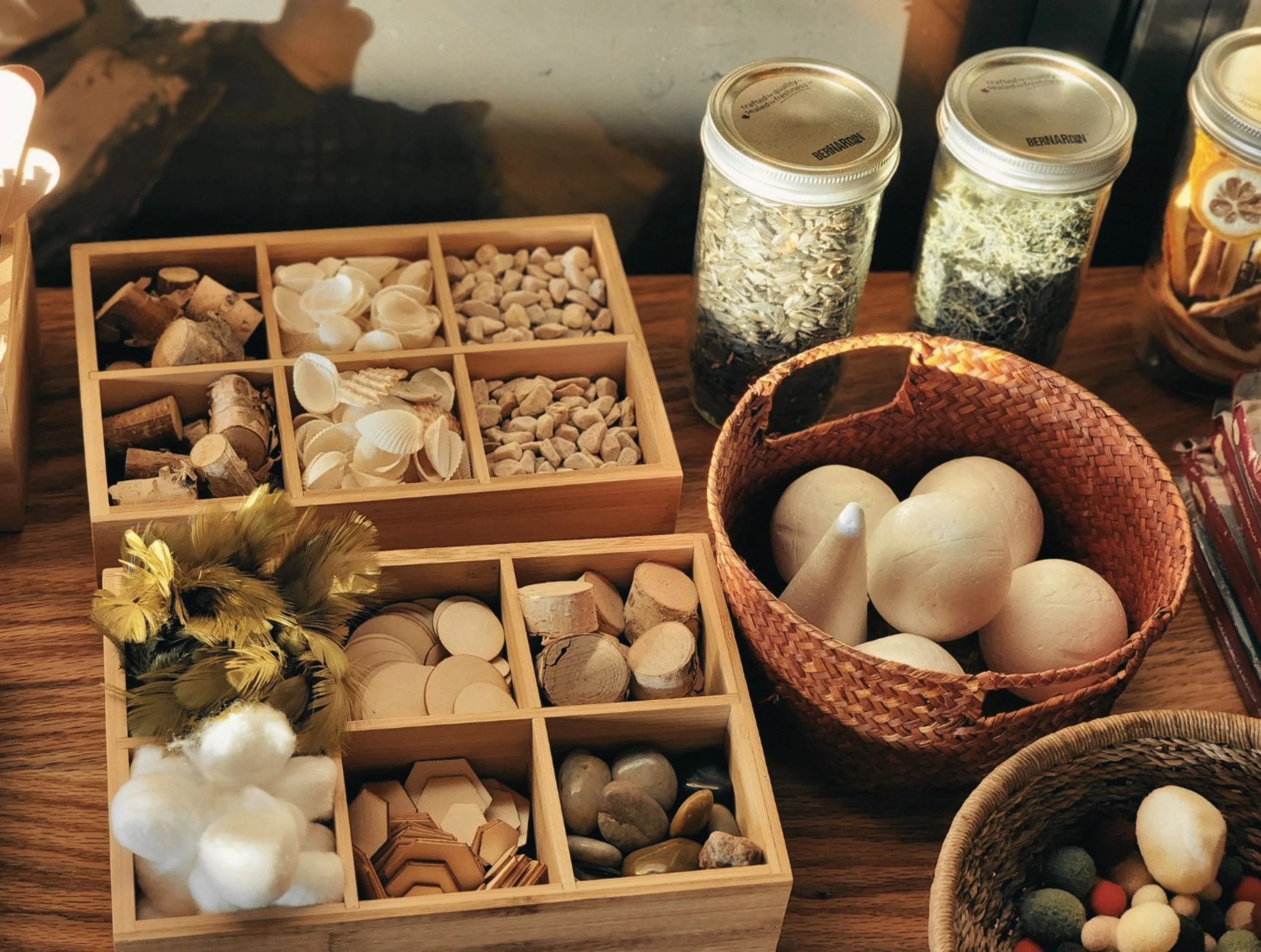Disposition d'éléments décoratifs en bois, plumes, pierres, coquillages, boules en coton, et bocaux contenant des herbes et des fleurs séchées sur une table en bois.