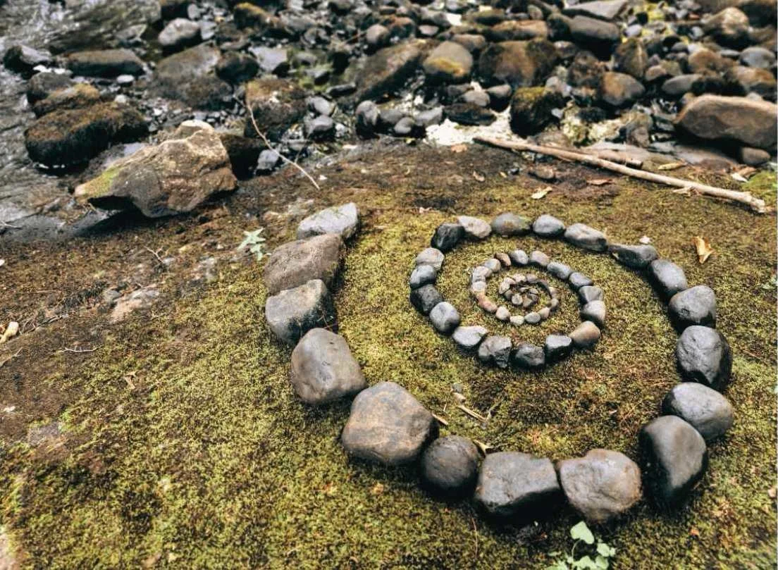 Des pierres disposées en spirale sur un sol recouvert de mousse, près d'une rivière ou d'une zone rocheuse.