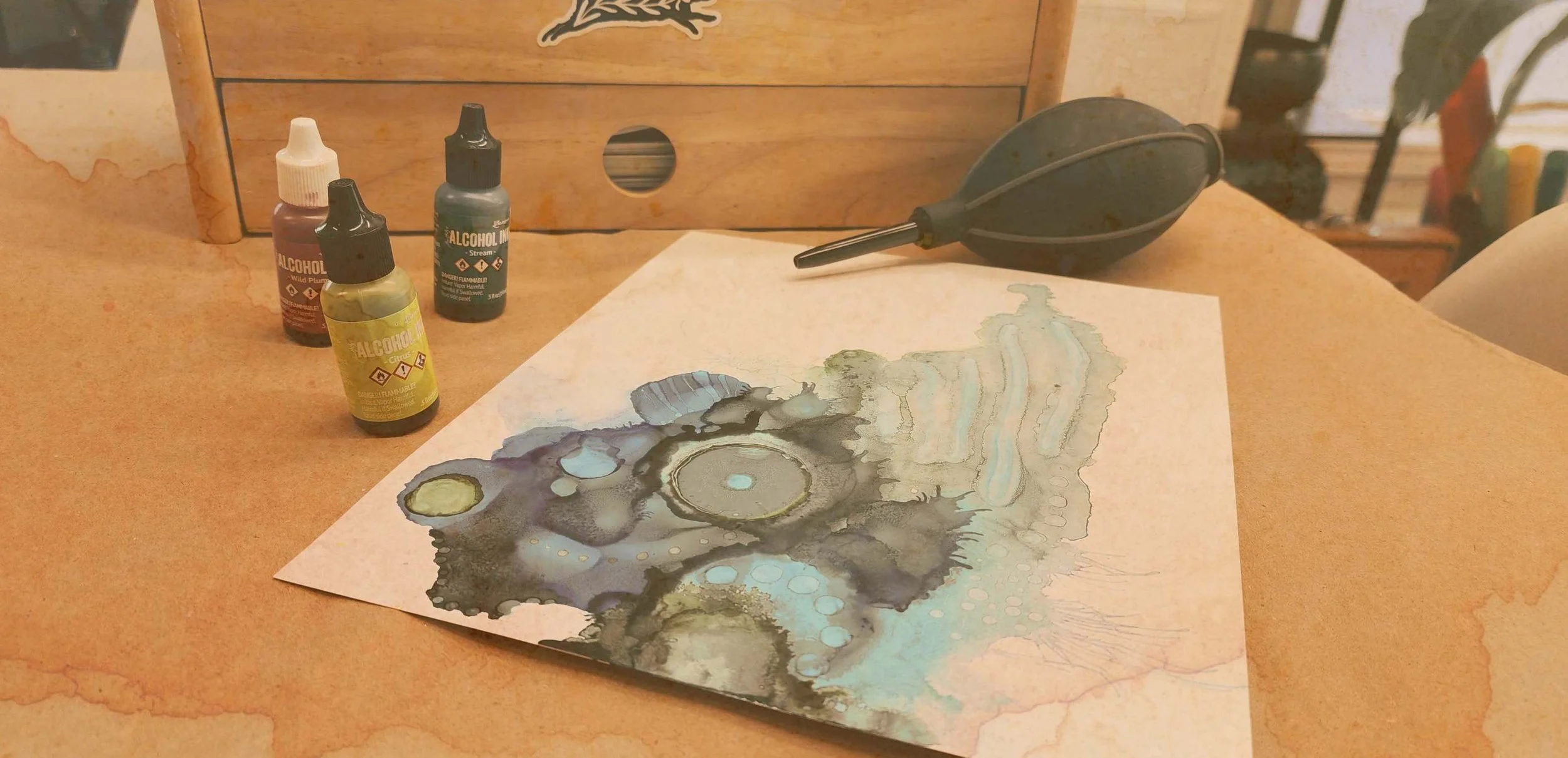 Une œuvre d'art abstraite créée avec des encres, posée sur une table en bois, avec des bouteilles de liquides colorés et un souffleur en caoutchouc à côté.