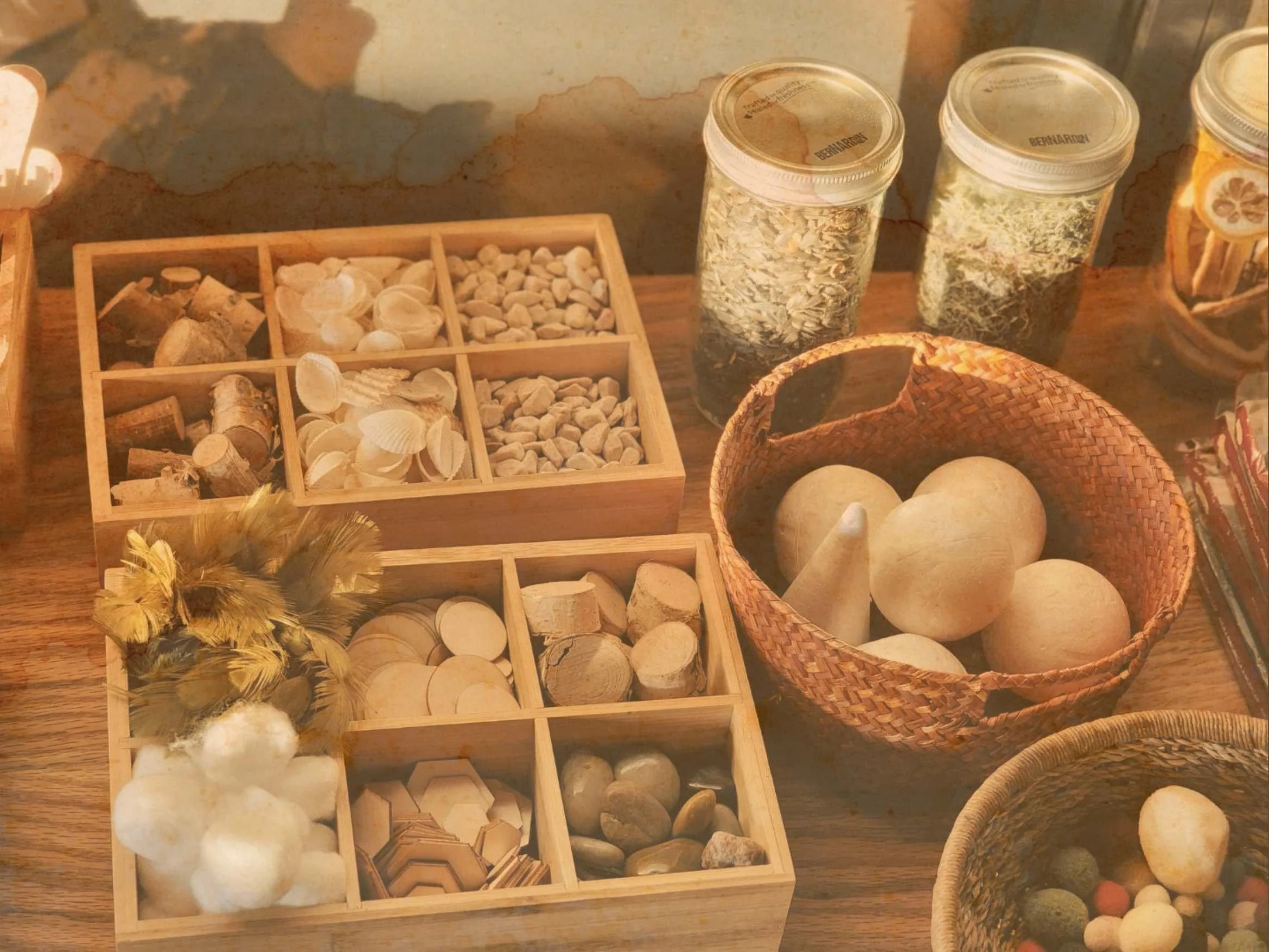 Différents contenants en bois et en verre remplis de diverses pierres, coquillages, coquines, et autres matériaux naturels, avec un panier en dessous contenant des pommes de terre et d'autres objets, sur une surface en bois.
