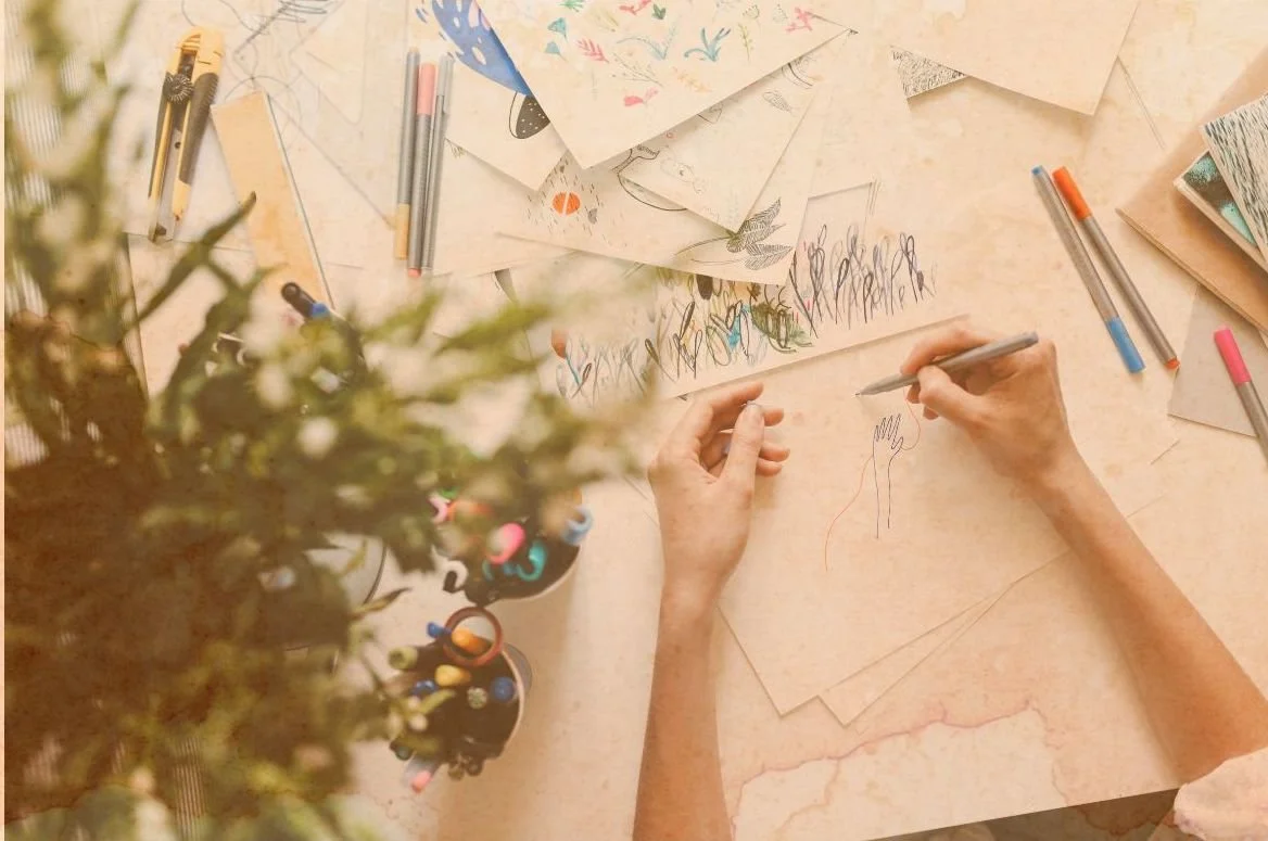 Une personne qui dessine à la main sur un papier en utilisant un stylo, entourée de plusieurs feuilles de papier avec des dessins et des notes, des stylos colorés et un pot de crayons, avec une plante floue en premier plan.