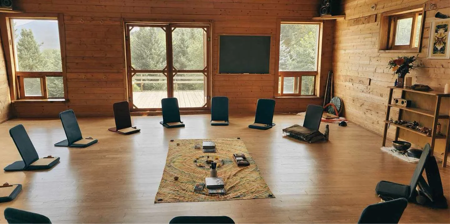 Retraite d'éco-art-thérapie, chaises disposées en cercle, tapis de sol, livres et objets ronds sur un tapis au centre, murs en bois, grandes fenêtres laissant entrer la lumière naturelle, vue sur la nature extérieure.